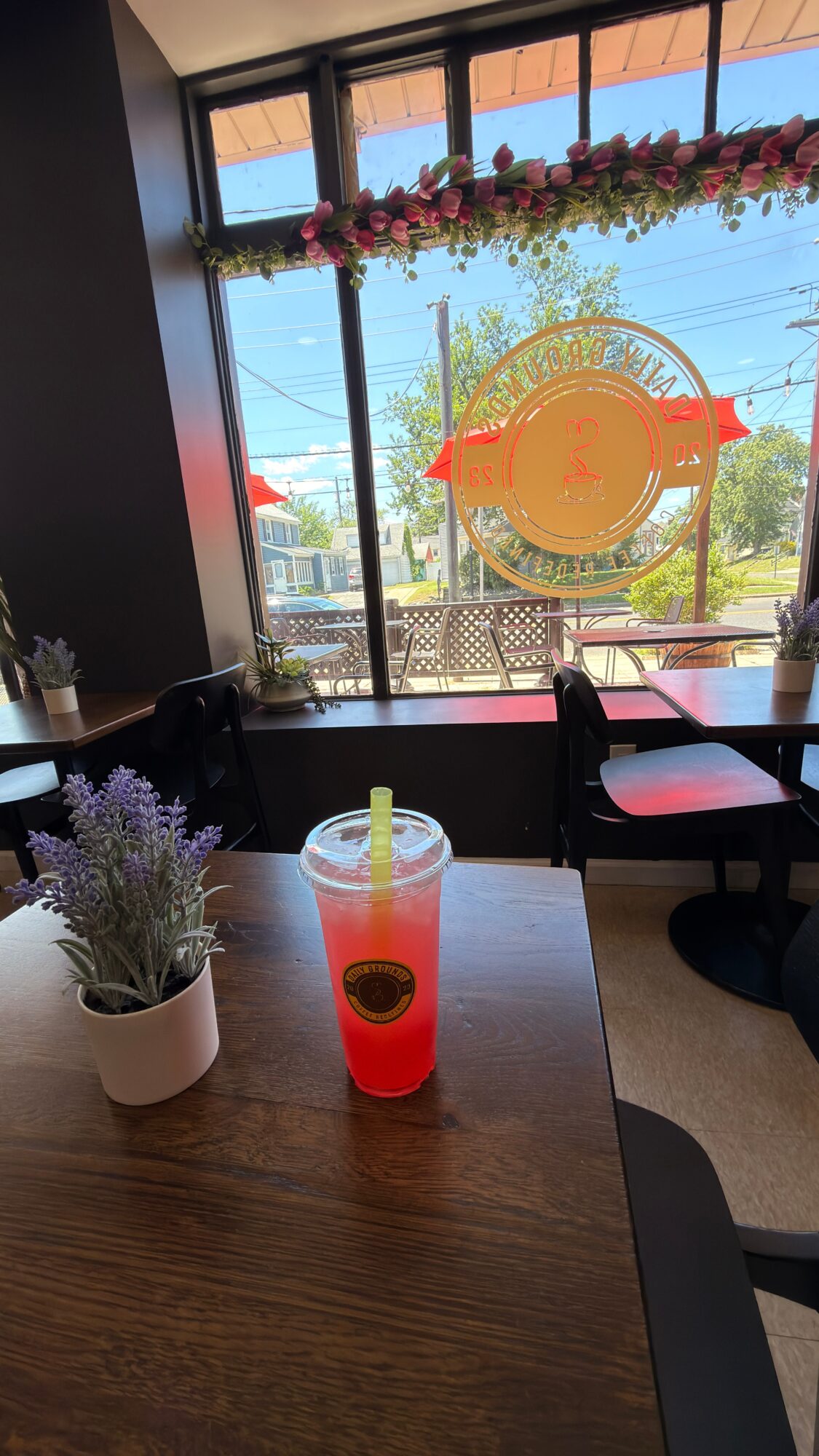 Pink drink with a straw on a table, potted lavender plant nearby, window with floral decoration outside, and a logo on the window.