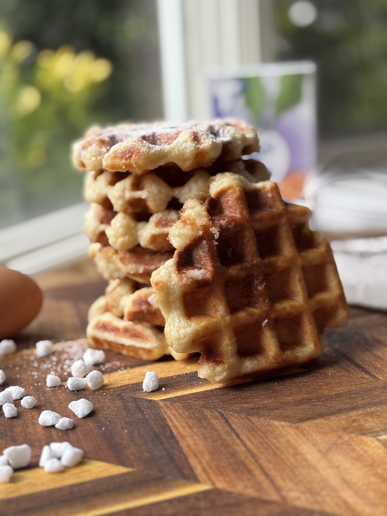 Stack of four waffles on a wooden surface with scattered sugar crystals, a mug, and a window in the background.