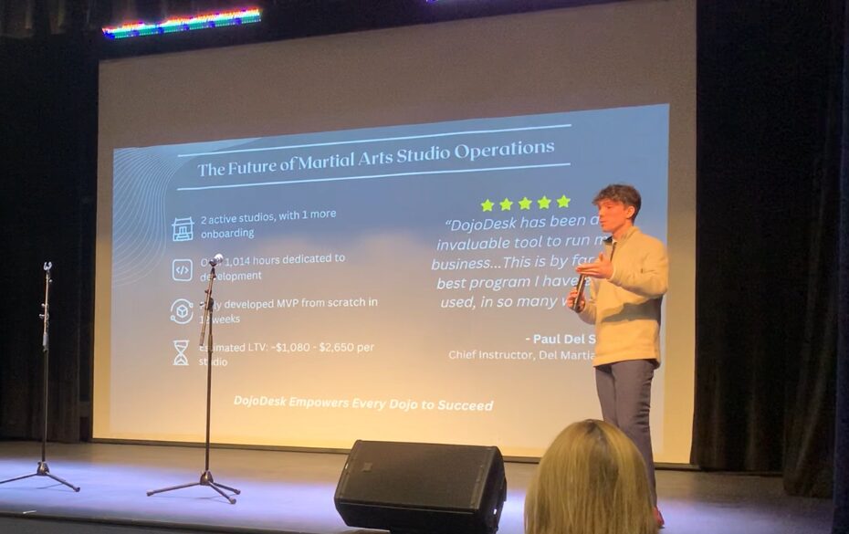 Person presenting on stage with a large screen displaying a presentation slide, audience visible in foreground.