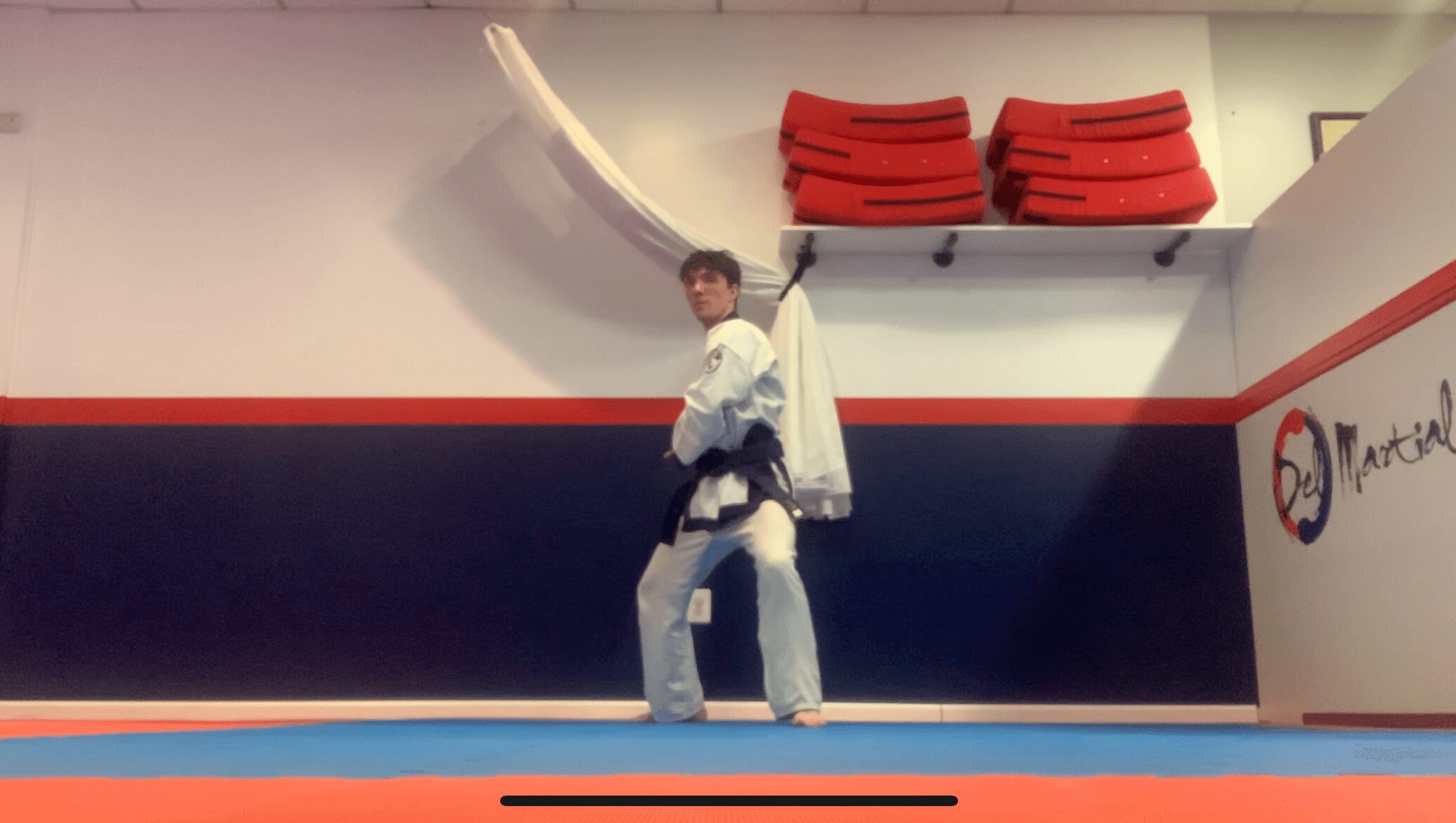 Martial artist in a white uniform and black belt standing on a mat in a dojo, with red mats stacked on a shelf above.