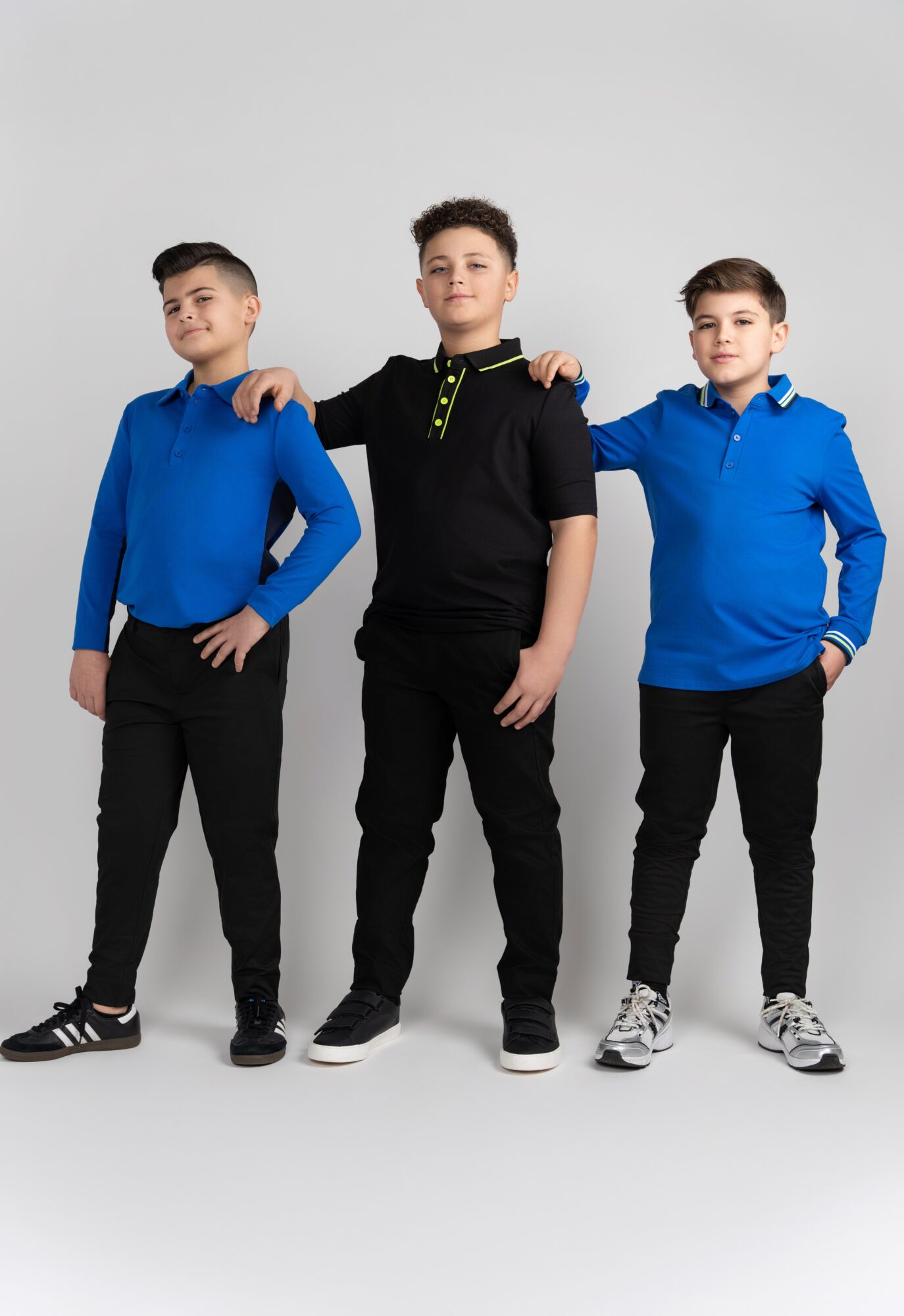 Three boys standing together, each with one arm on another's shoulder, wearing blue and black clothing, against a plain background.
