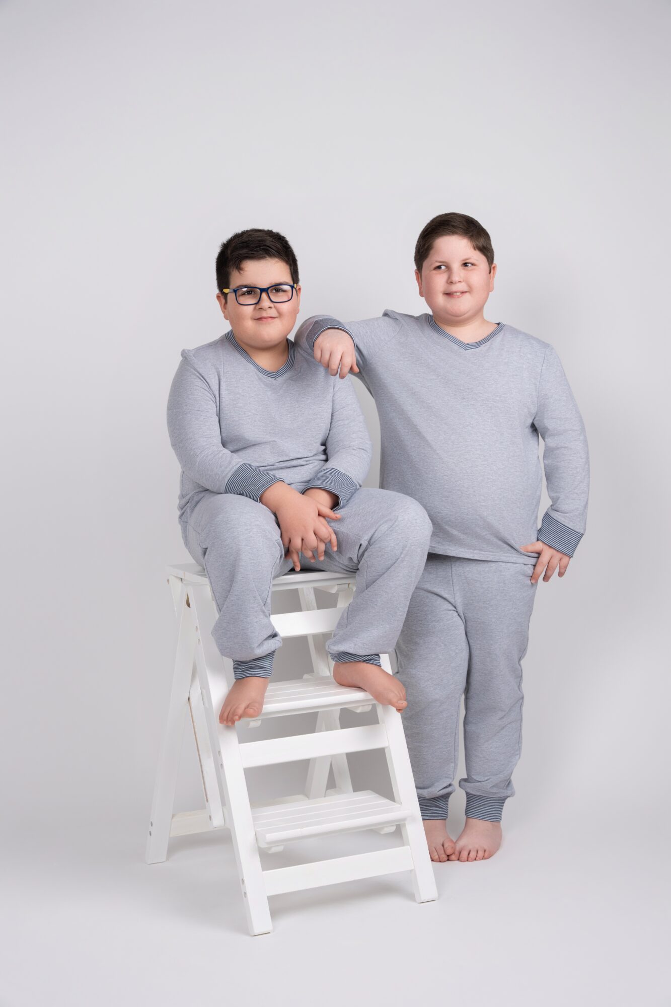 Two boys in gray pajamas, one sitting on a small white ladder and the other standing beside it, smiling.