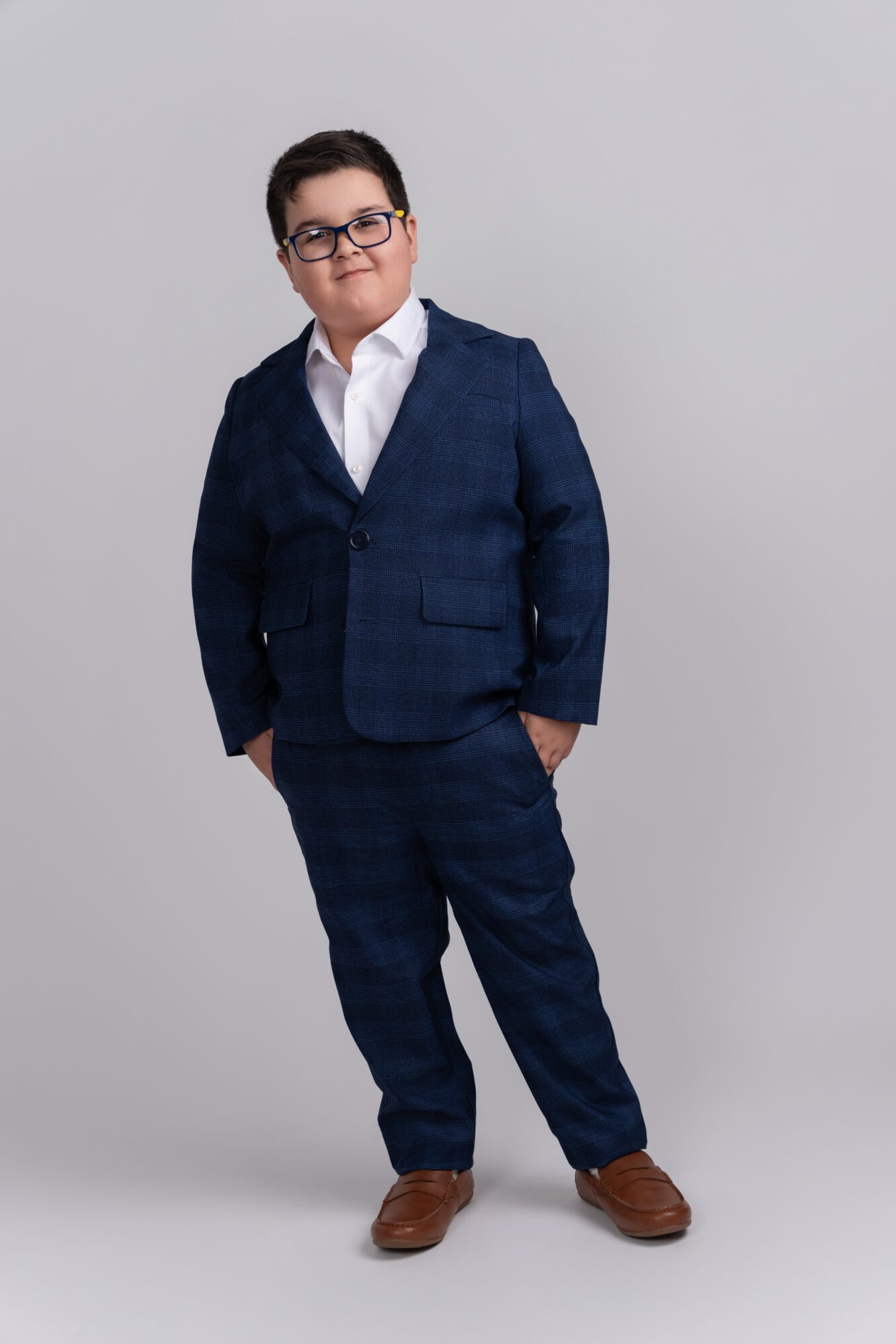 Young boy in a navy blue suit, white shirt, glasses, standing with hands in pockets, smiling slightly, against a plain gray background.