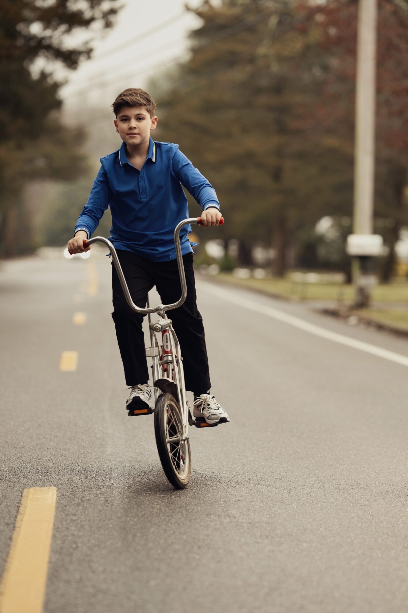 Boy riding a bicycle on a street with trees and houses in the background, wearing a blue jacket and black pants.