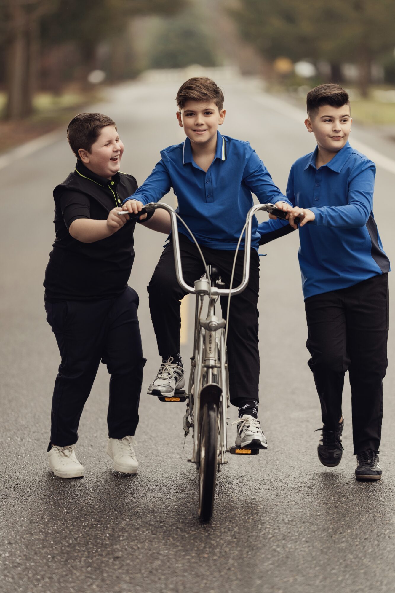 Three boys walking on a street, one riding a bicycle, smiling and holding the handlebars, with trees in the background.