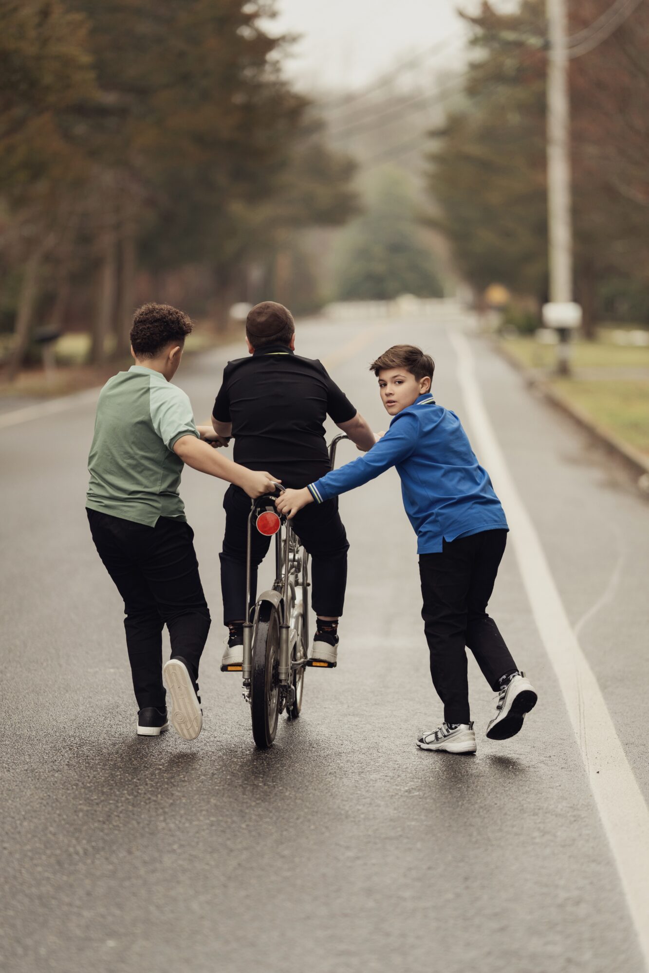 Three boys walk on a road, one pushes a bicycle while the others hold its handlebars, trees and streetlights in background.