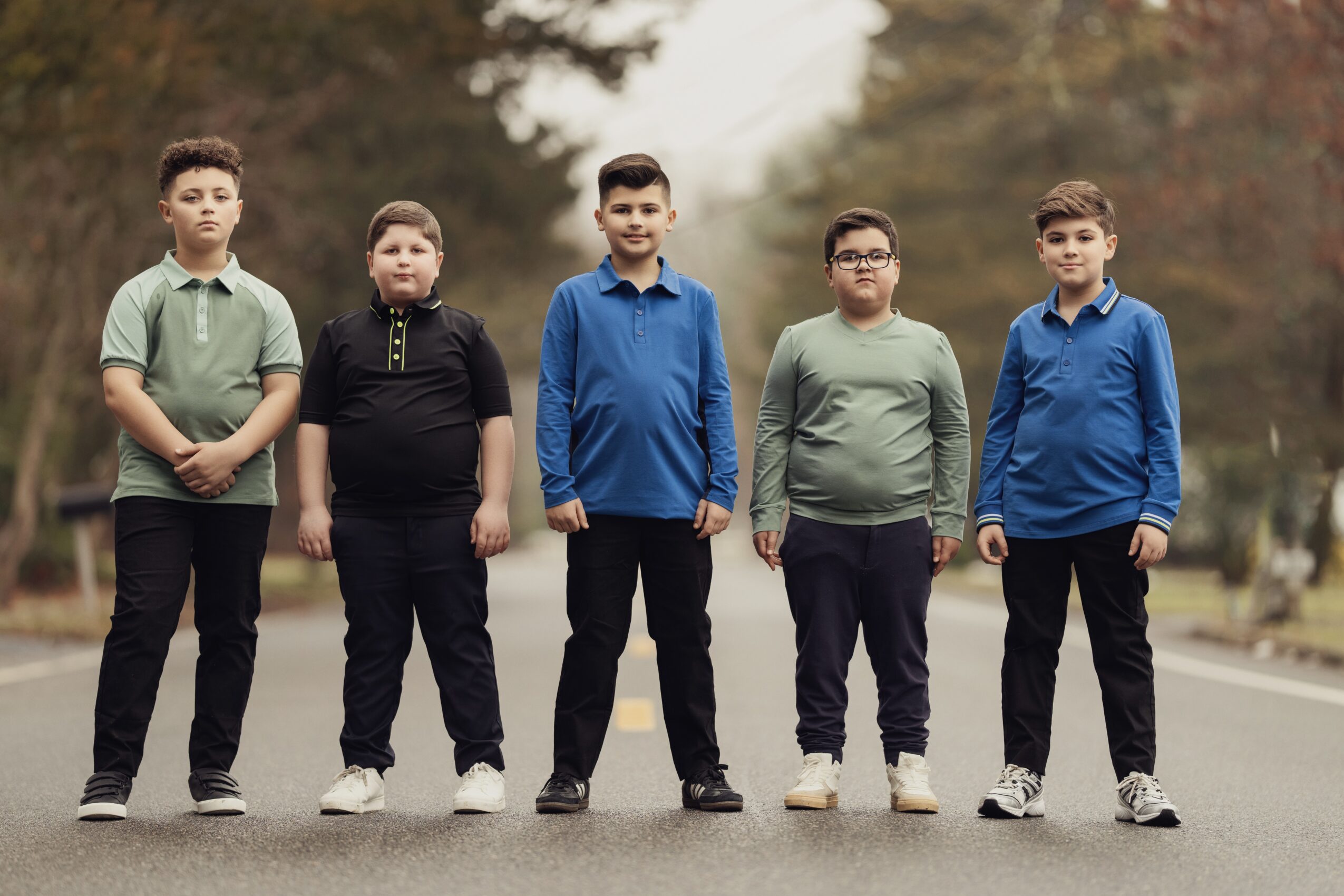 Five boys standing outdoors on a sidewalk with trees in the background, wearing casual clothing and looking at the camera.