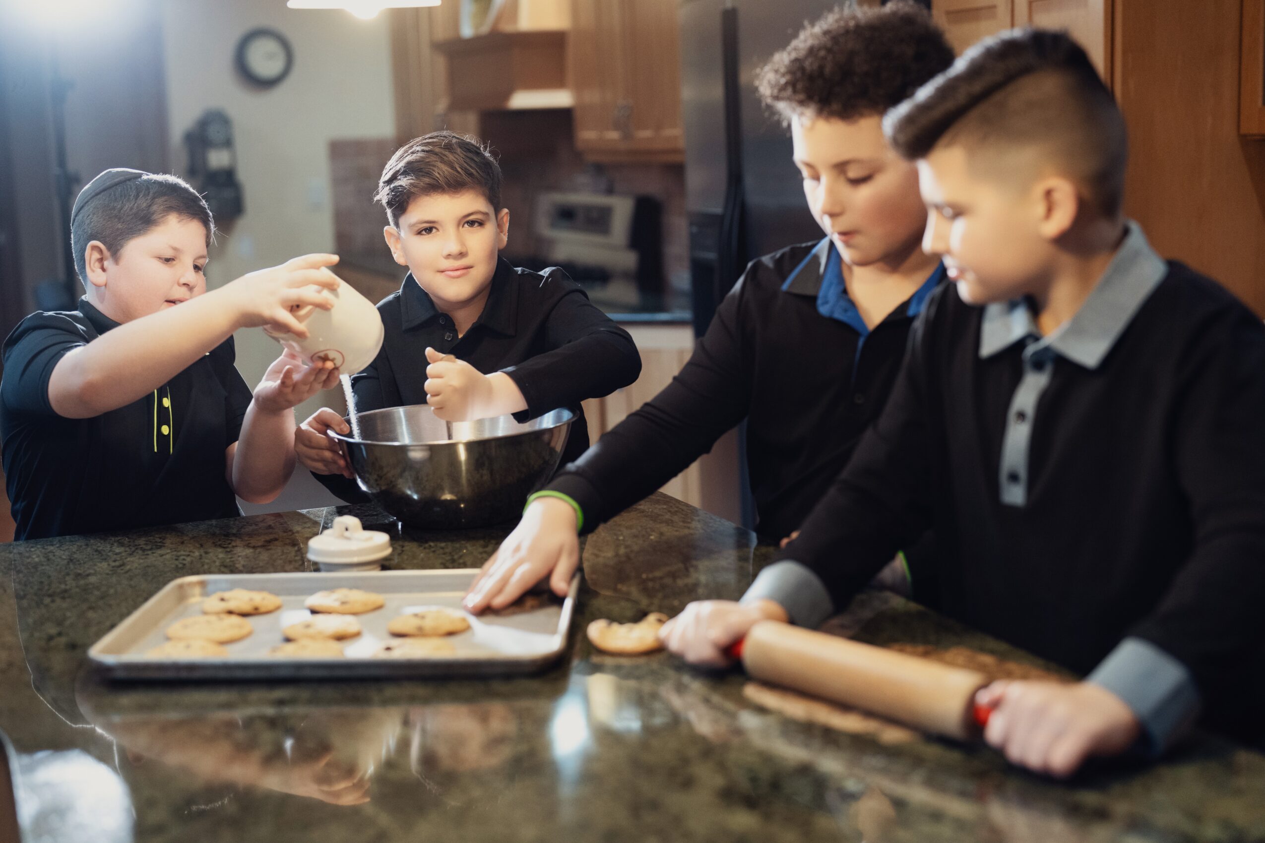 Four children baking cookies in a kitchen, one using a rolling pin, others handling dough and a baking tray.