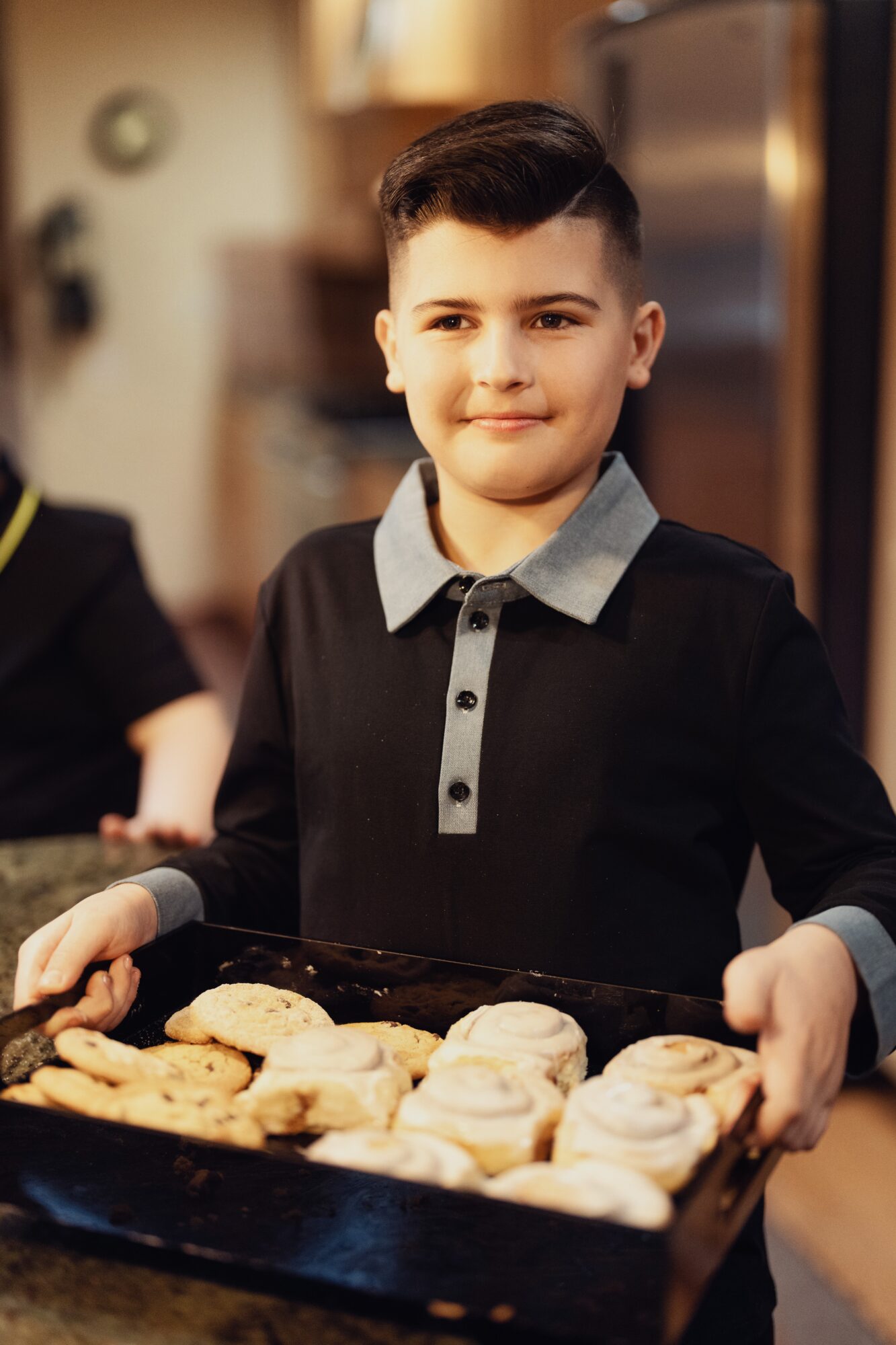 Young boy with short dark hair and a gray collared shirt holding a tray of baked cookies, smiling.