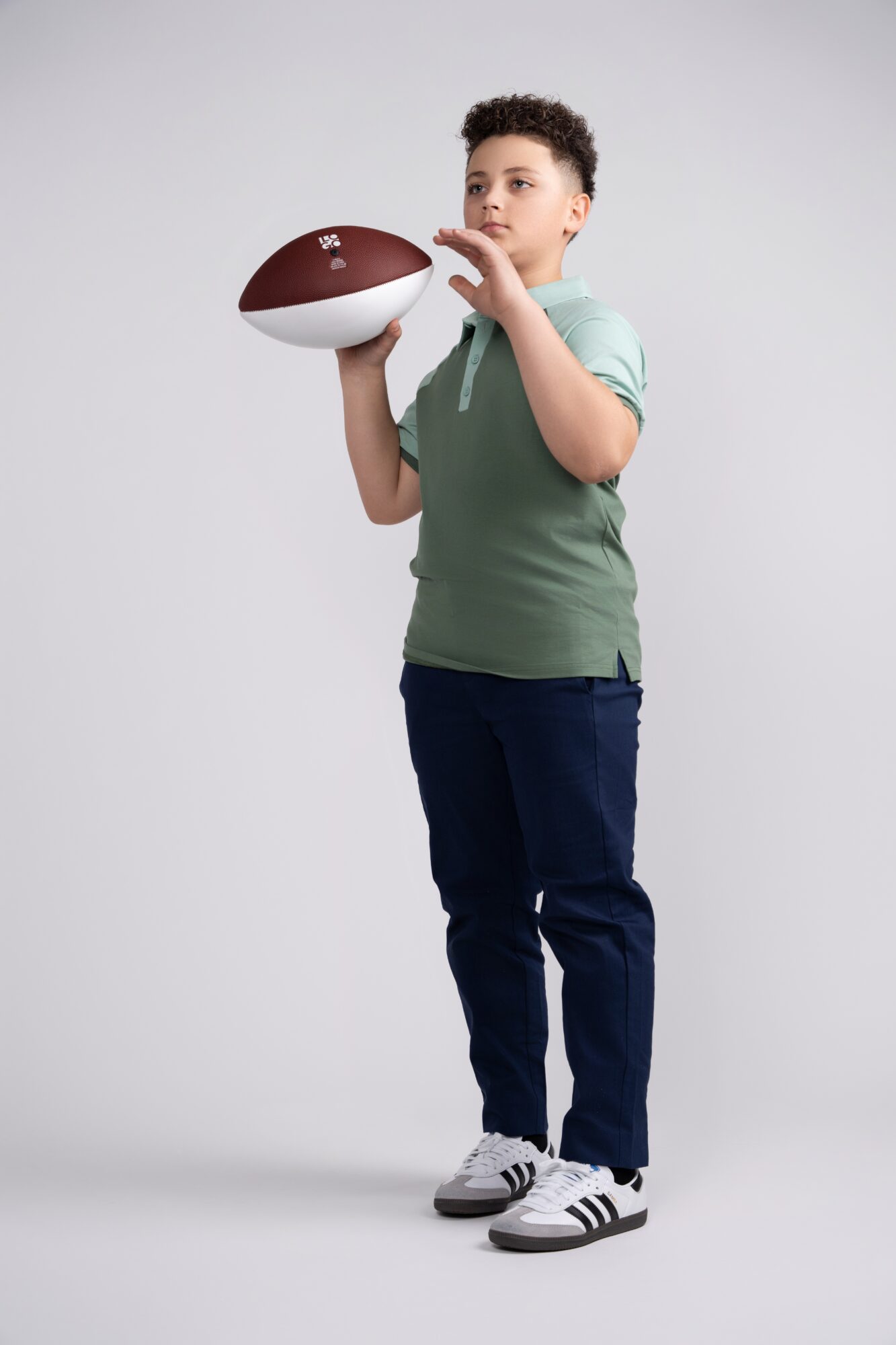 Young boy holding a football, standing against a plain background, wearing a green shirt, dark pants, and sneakers.