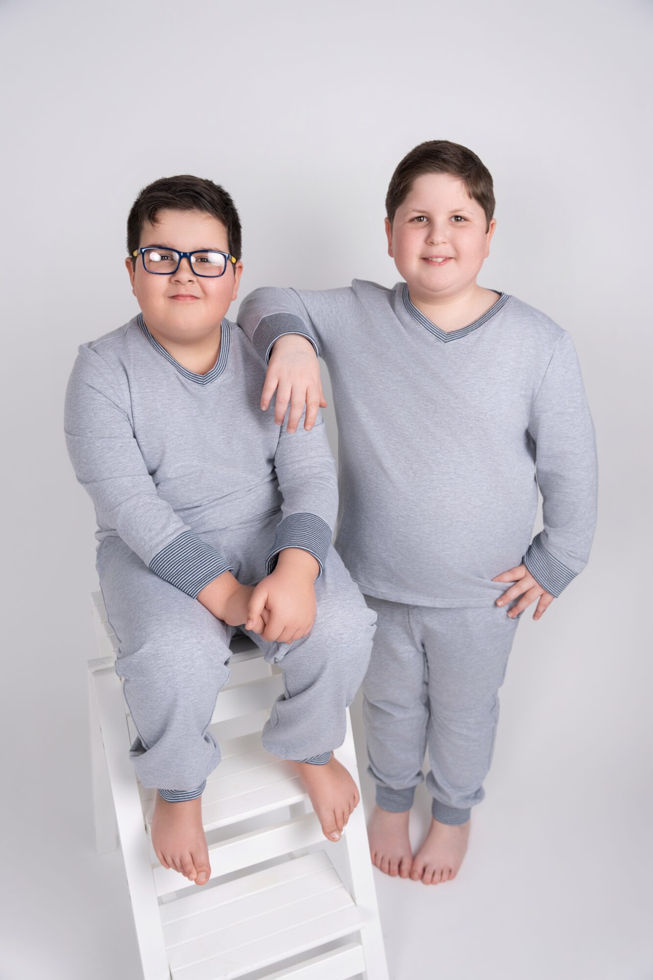 Two children in gray outfits pose against a plain background, one sitting on a stool and the other standing with hand on hip.