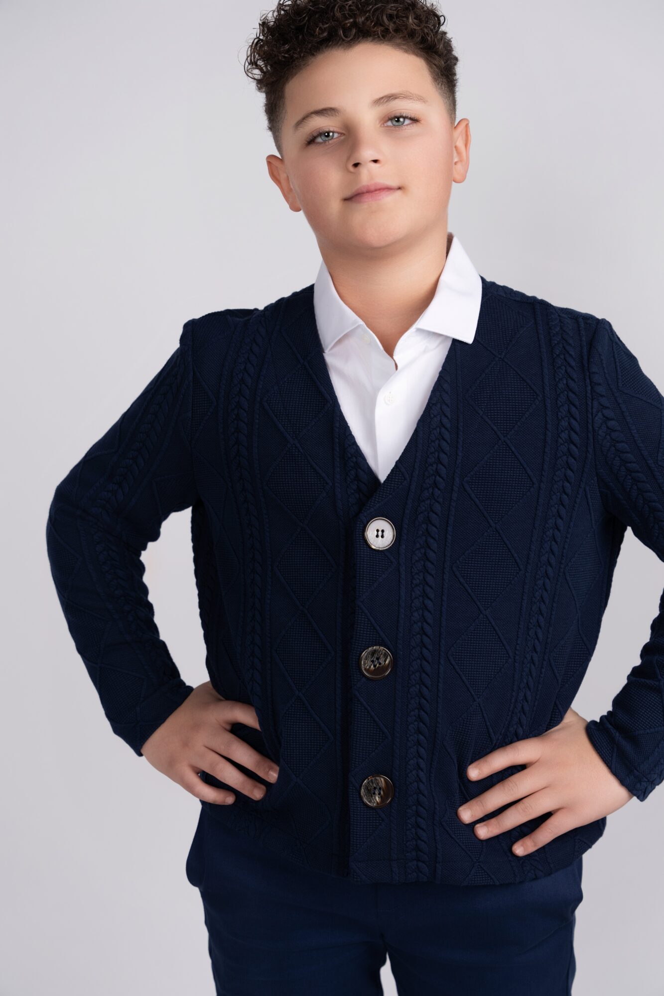 Young boy with short curly hair wearing a white shirt and navy blue cardigan, standing with hands on hips.