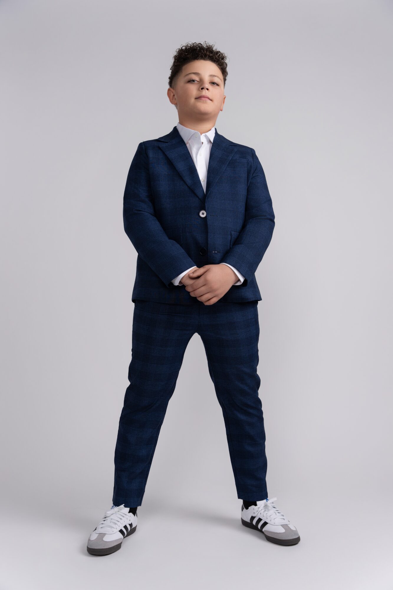 Young boy in a navy suit with white shirt, standing confidently with hands clasped, against a plain light background.