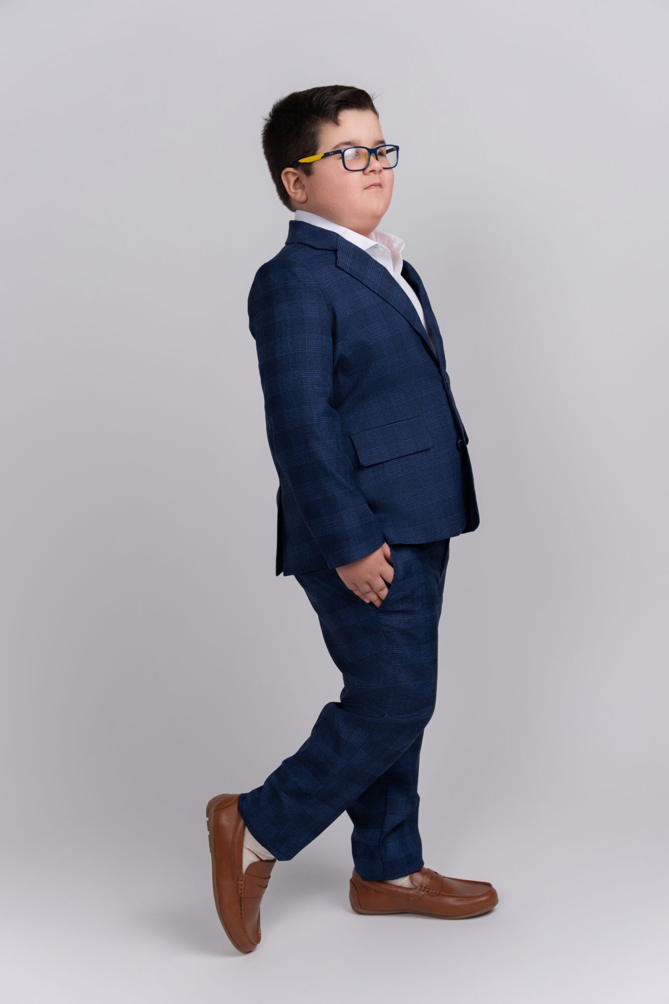 Boy in a suit and glasses standing sideways, facing right, with a neutral background.