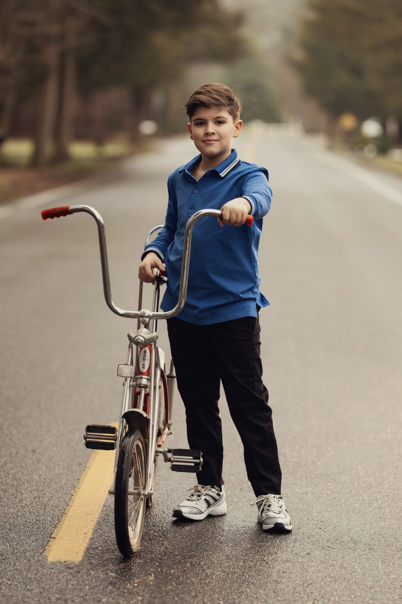 Boy standing on street holding bicycle, wearing blue jacket and black pants, with trees in background.