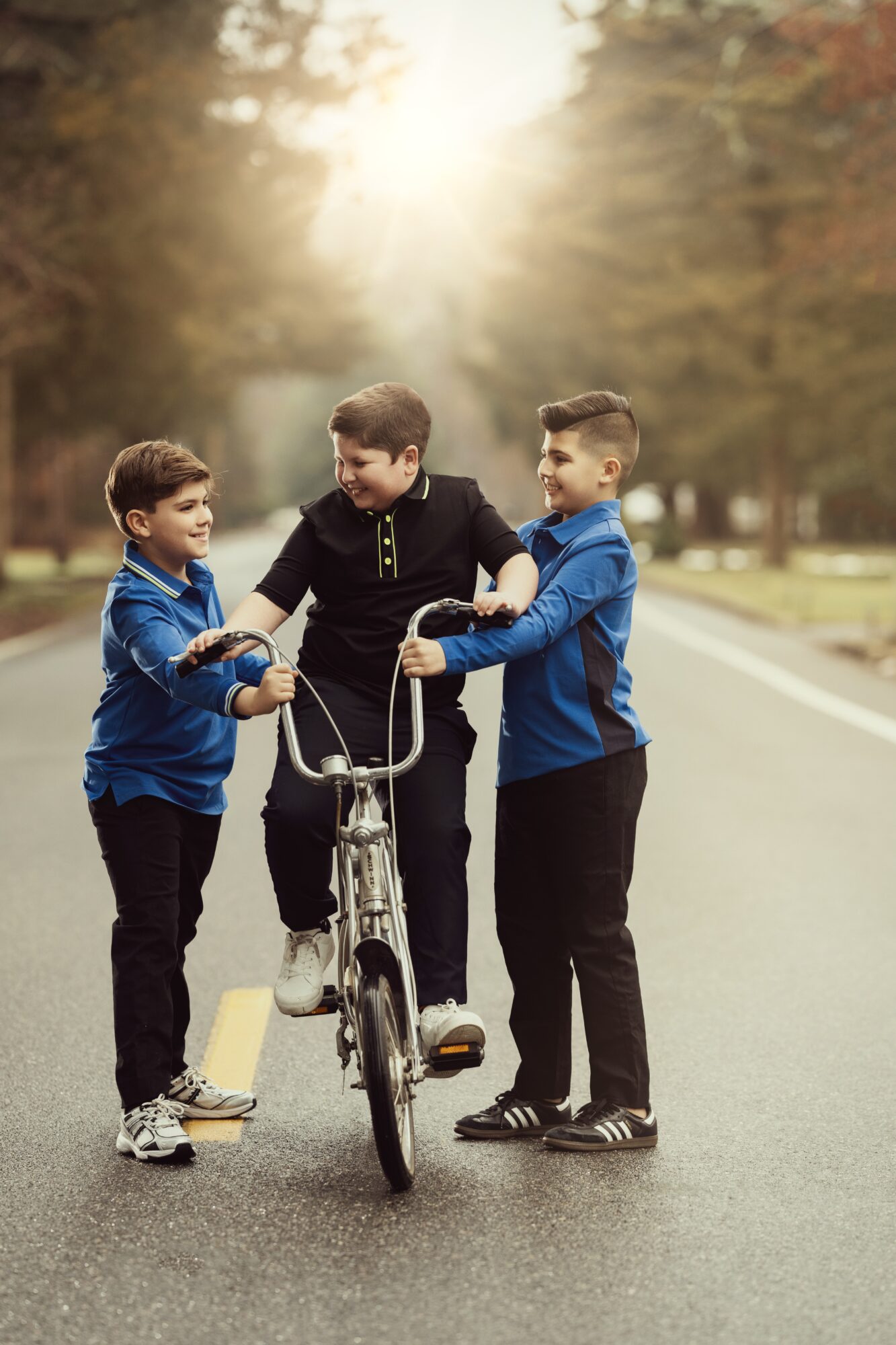 Three boys standing on a street, one with a bicycle, holding the handlebars, with trees and sunlight in the background.
