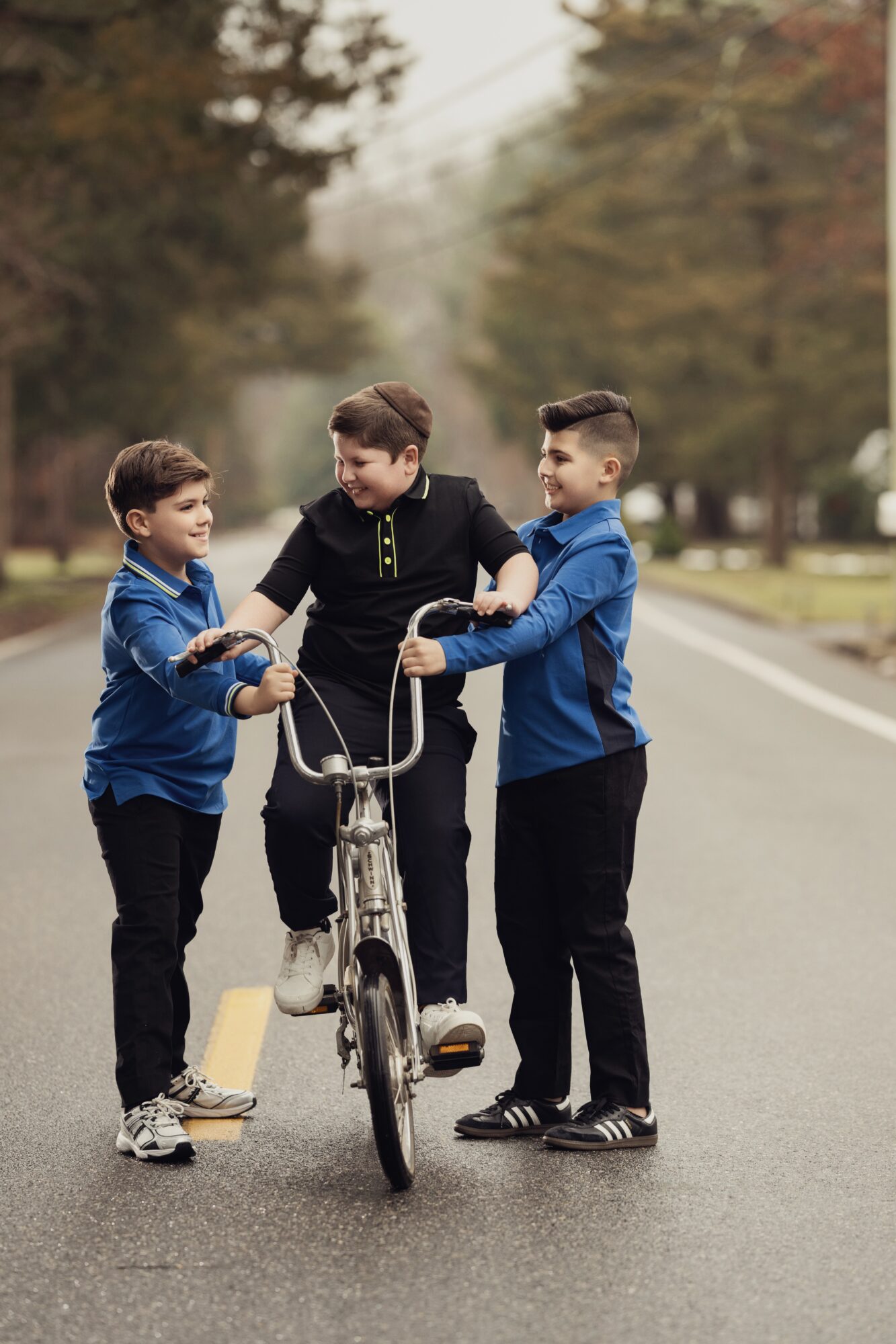 Three boys stand on a street, with one holding a bicycle, engaging in conversation outdoors during daytime.