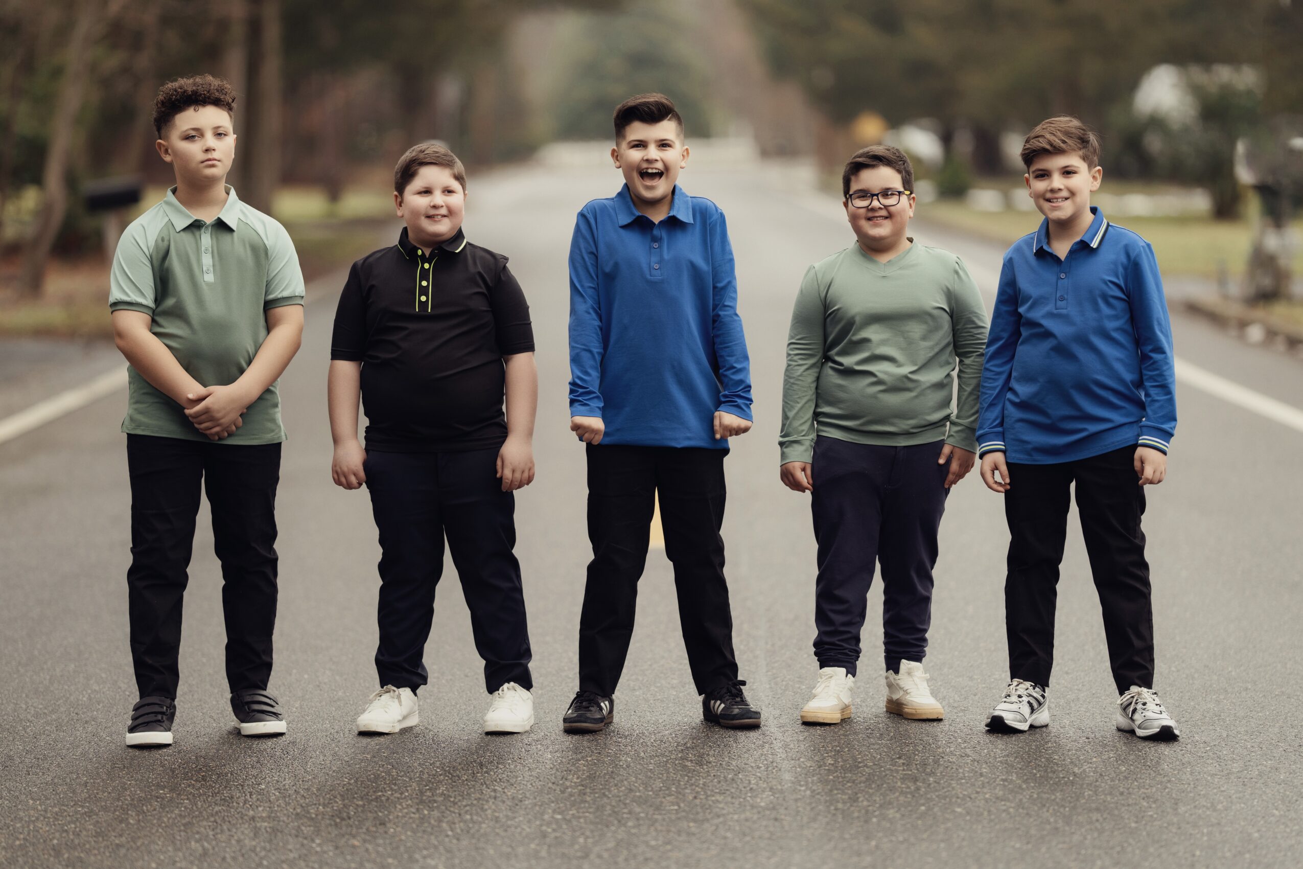 Five children stand in a row outdoors on a paved path, smiling and facing forward, with trees in the background.