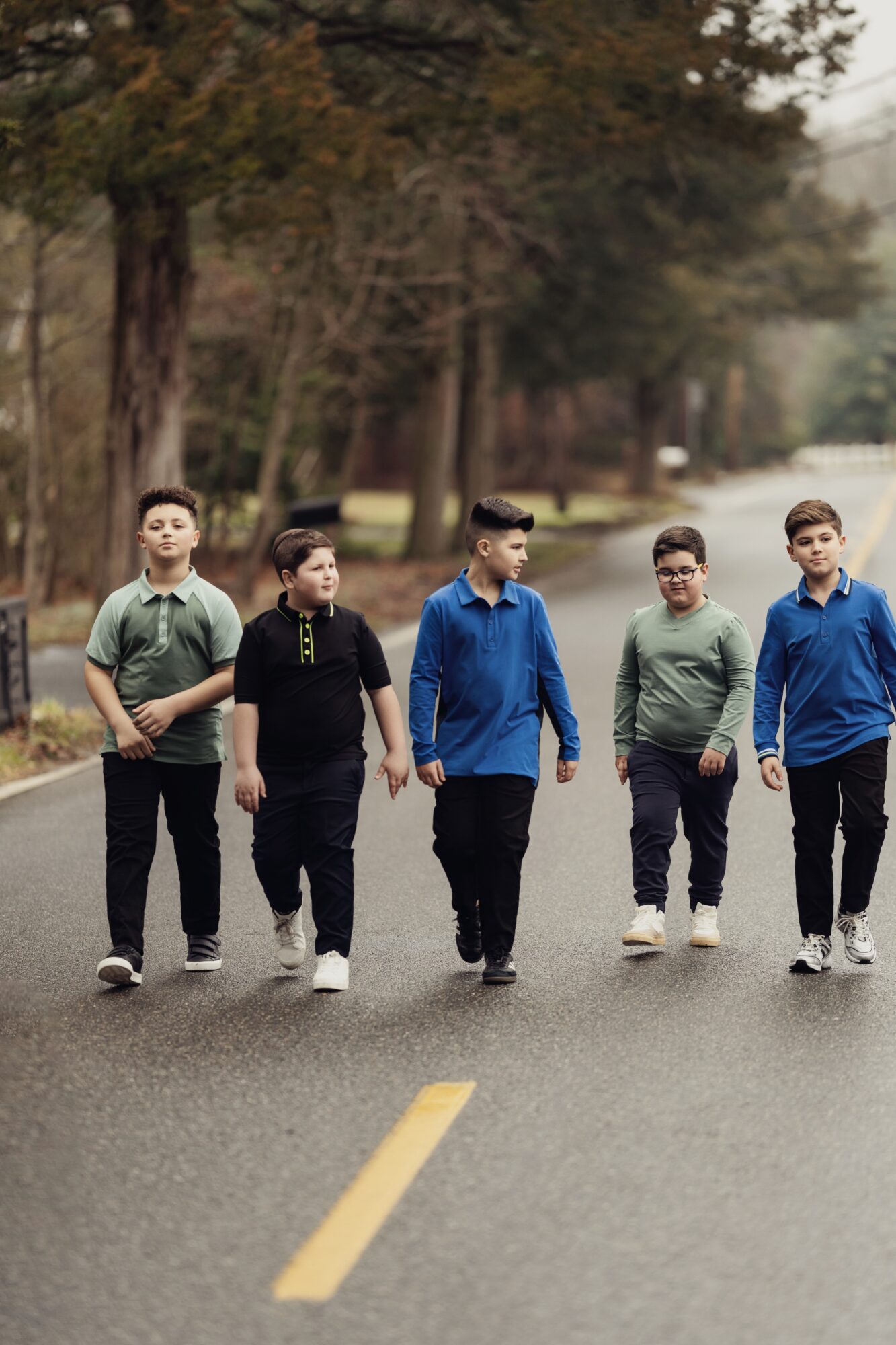 Five children walking on a road with trees in the background, wearing casual clothing, some with hands in pockets.