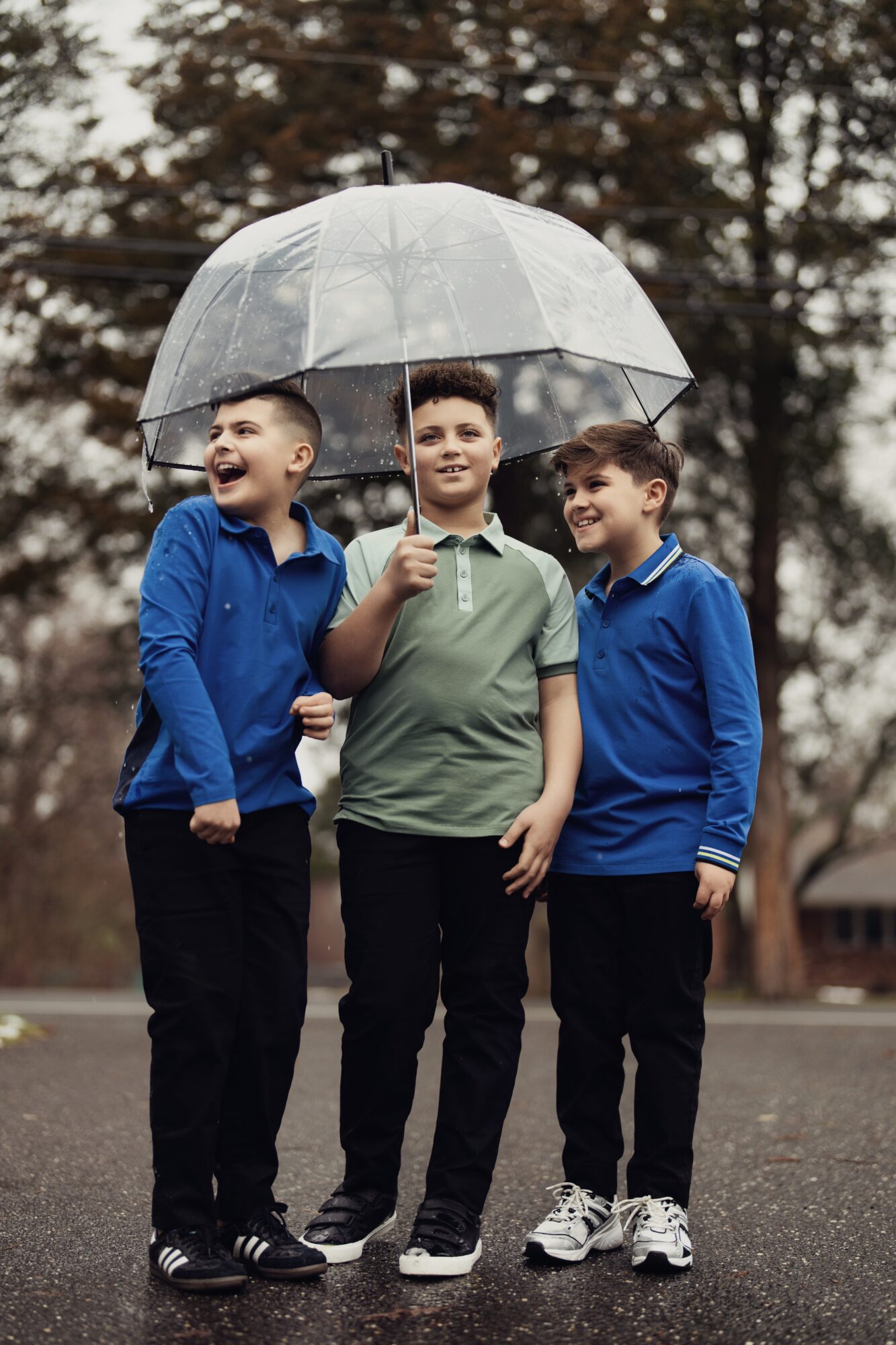 Three boys standing outdoors under a large transparent umbrella, smiling and looking in different directions.