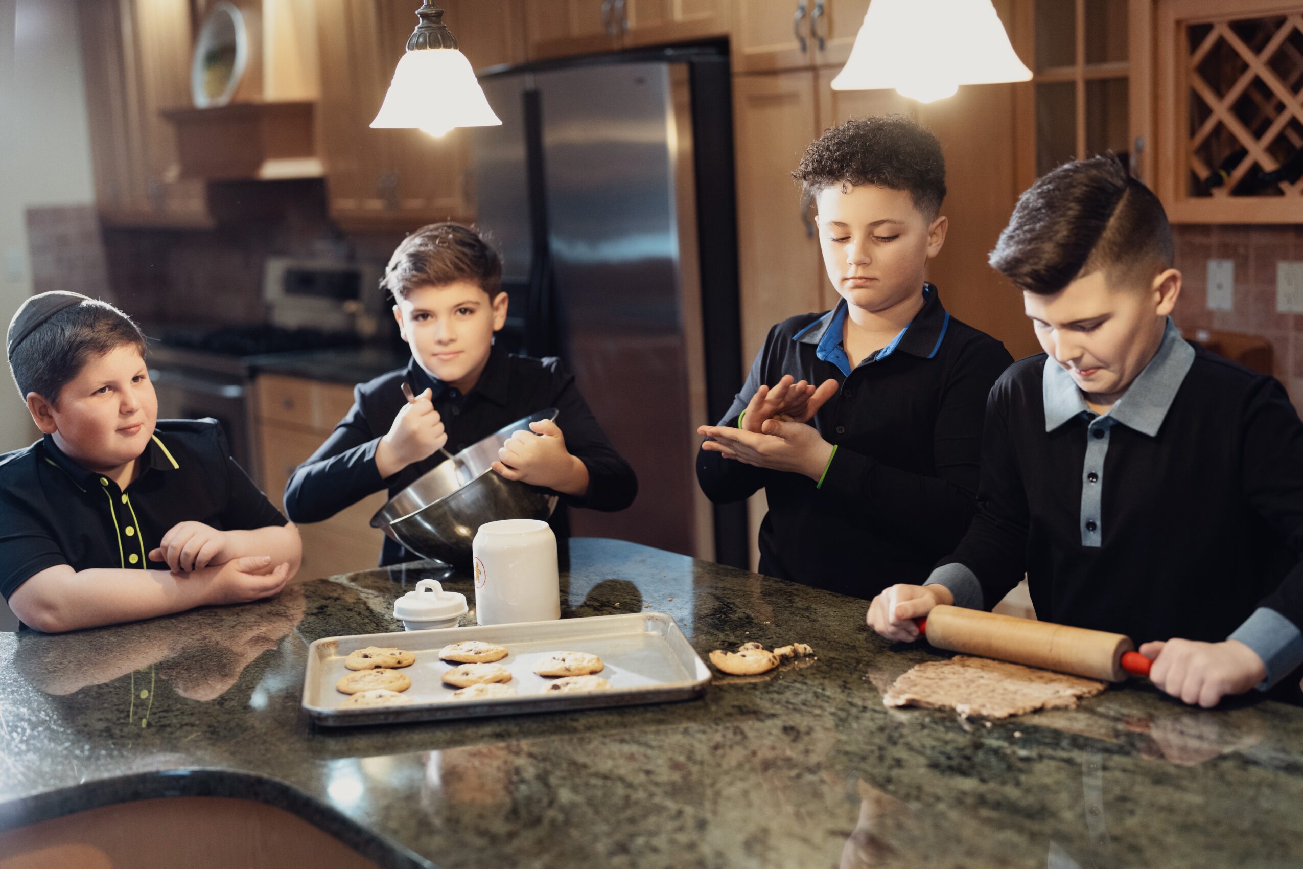 Four boys baking cookies in a kitchen, one rolling dough, others preparing ingredients, with a tray of cookies on the counter.