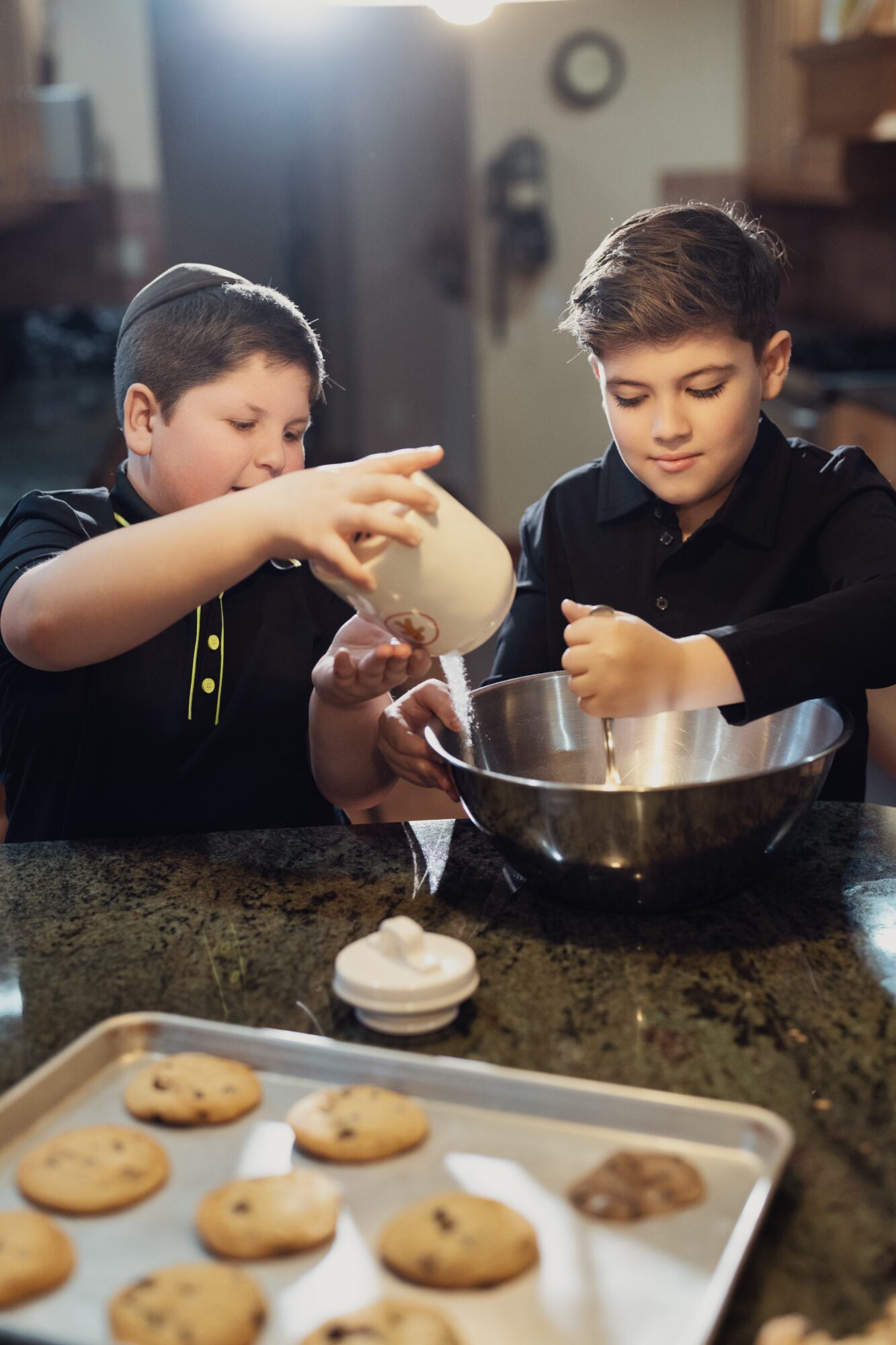Two boys baking cookies in a kitchen, pouring ingredients into a mixing bowl, with cookies on a tray in the foreground.