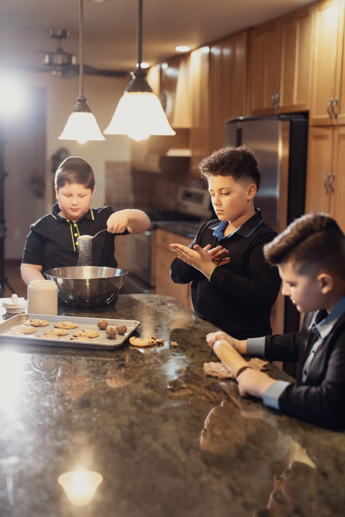 Three children in a kitchen preparing food, with a tray of cookies and a bowl on the counter.