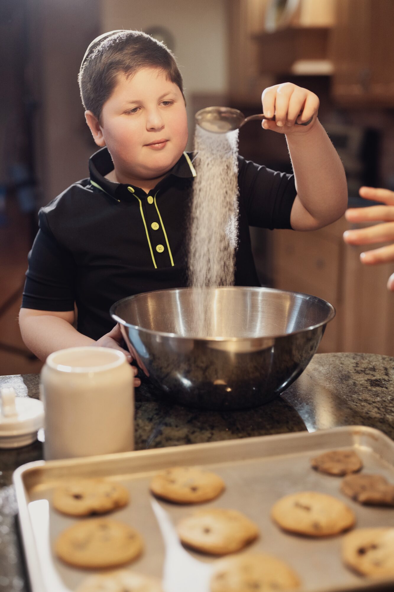 Child pouring sugar into a mixing bowl in a kitchen, cookies on a tray in foreground.