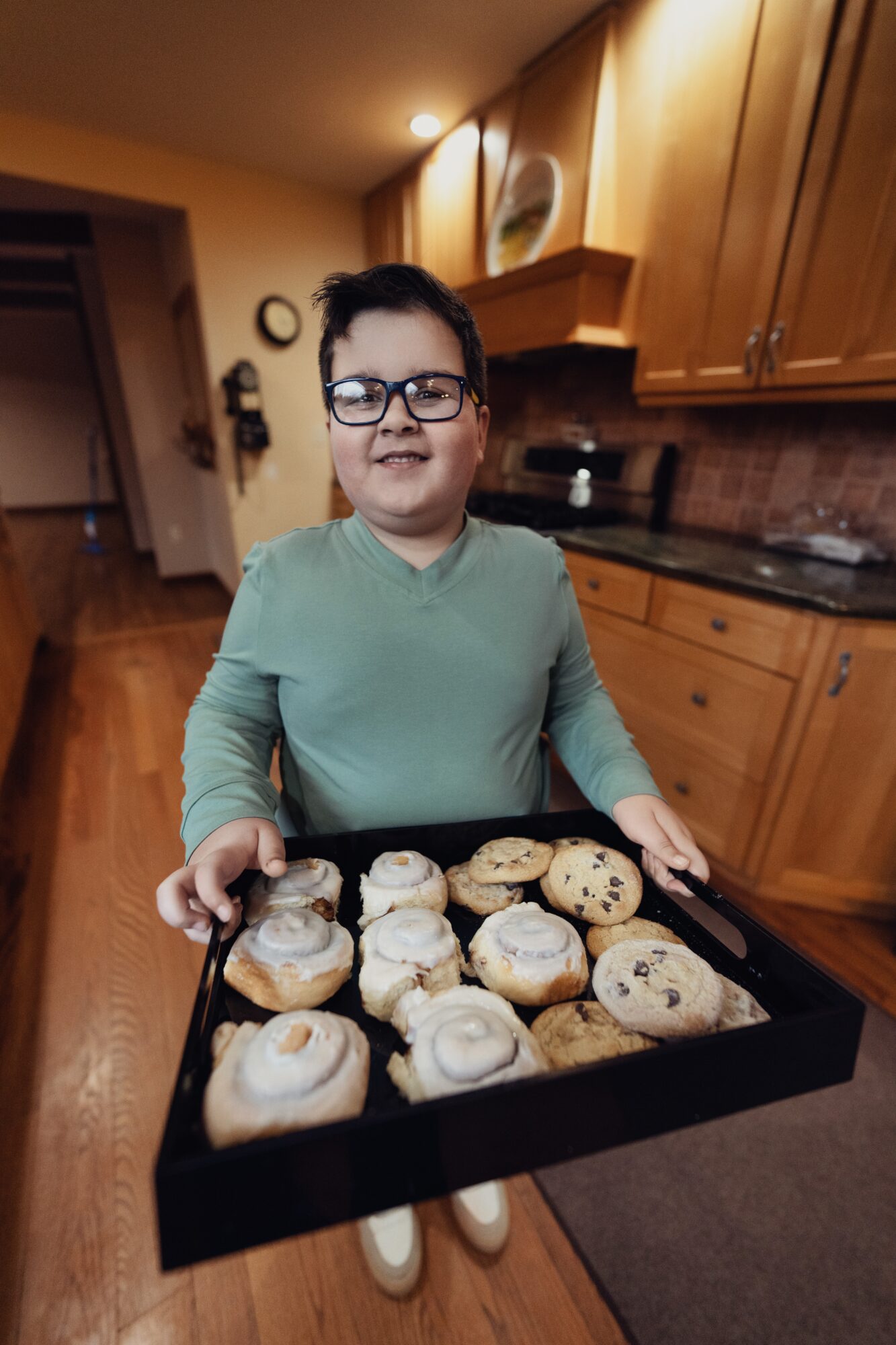 Child with glasses holding a tray of cookies in a kitchen, smiling, wooden cabinets in background.