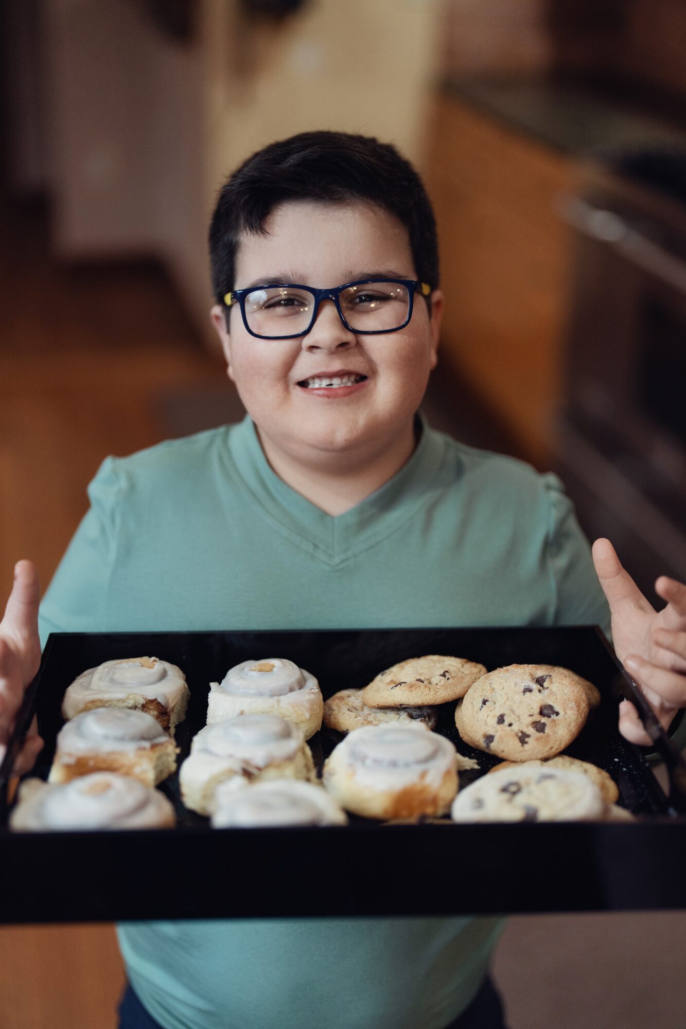 Child with glasses smiling, holding a tray of cookies, in a kitchen, wearing a green shirt.