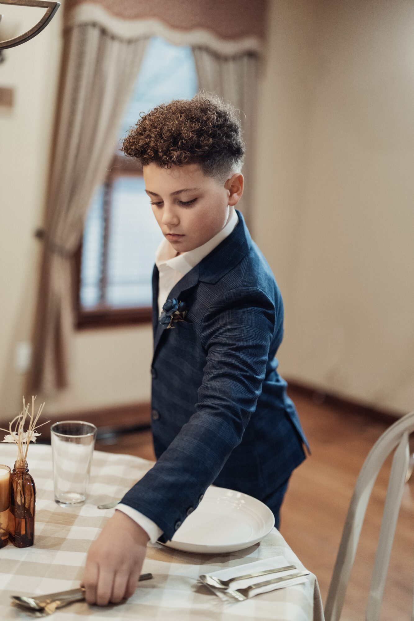 Young boy in a suit setting a table with a plate, glass, and utensils in a room with curtains and a window.