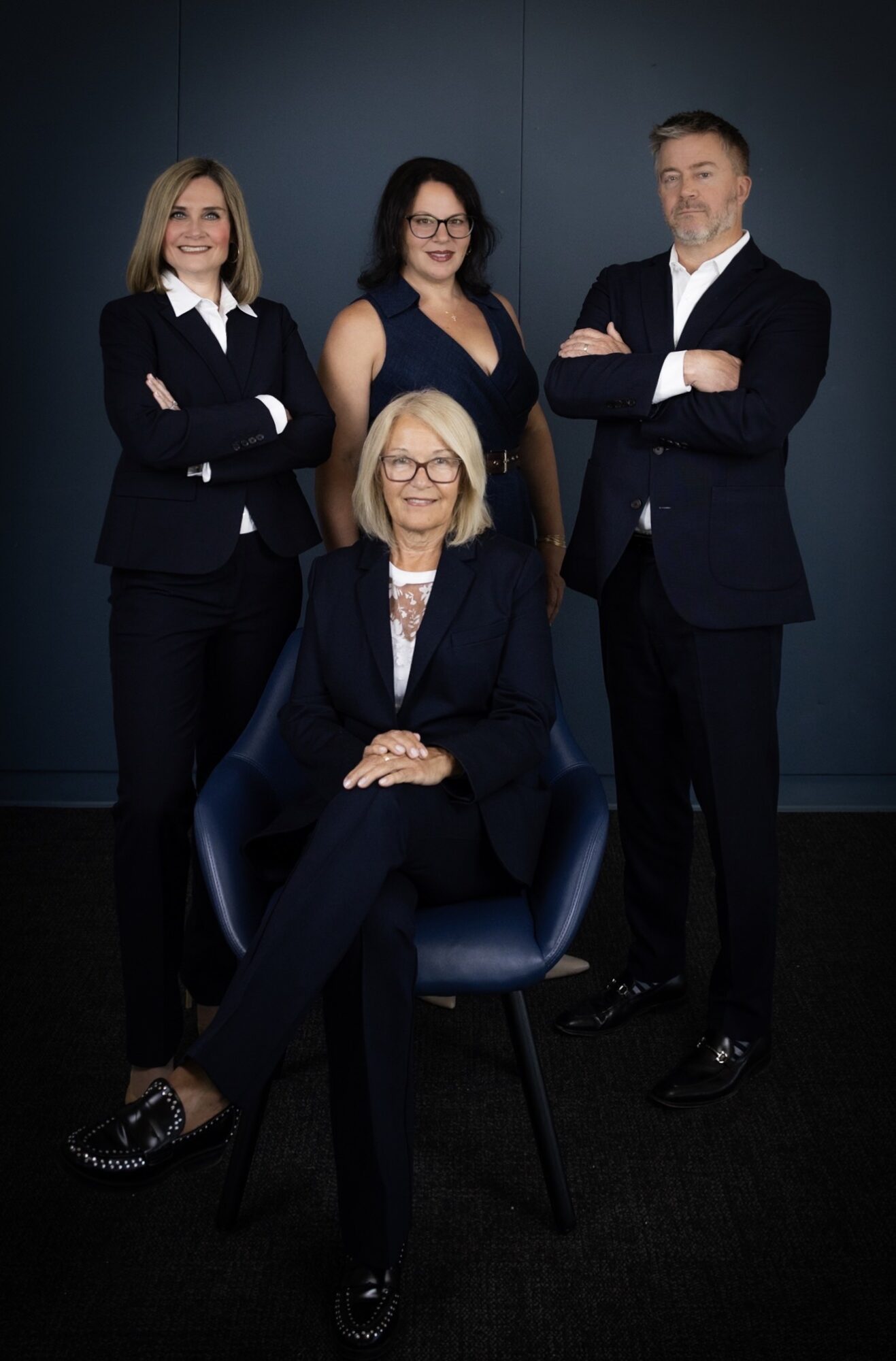 Four professionally dressed women pose against a dark background, one seated and three standing with arms crossed.