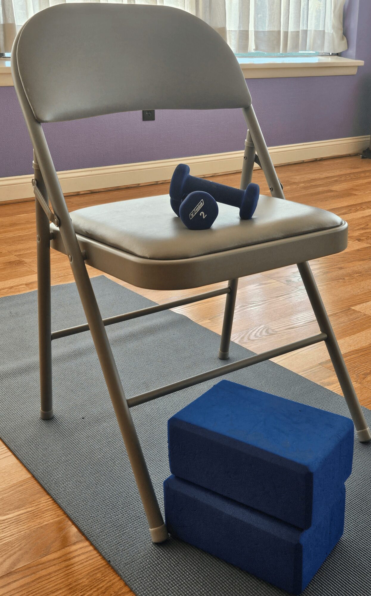 Folding chair with a pair of small dumbbells on the seat and two blue yoga blocks underneath, in a room with wooden floor and purple wall.