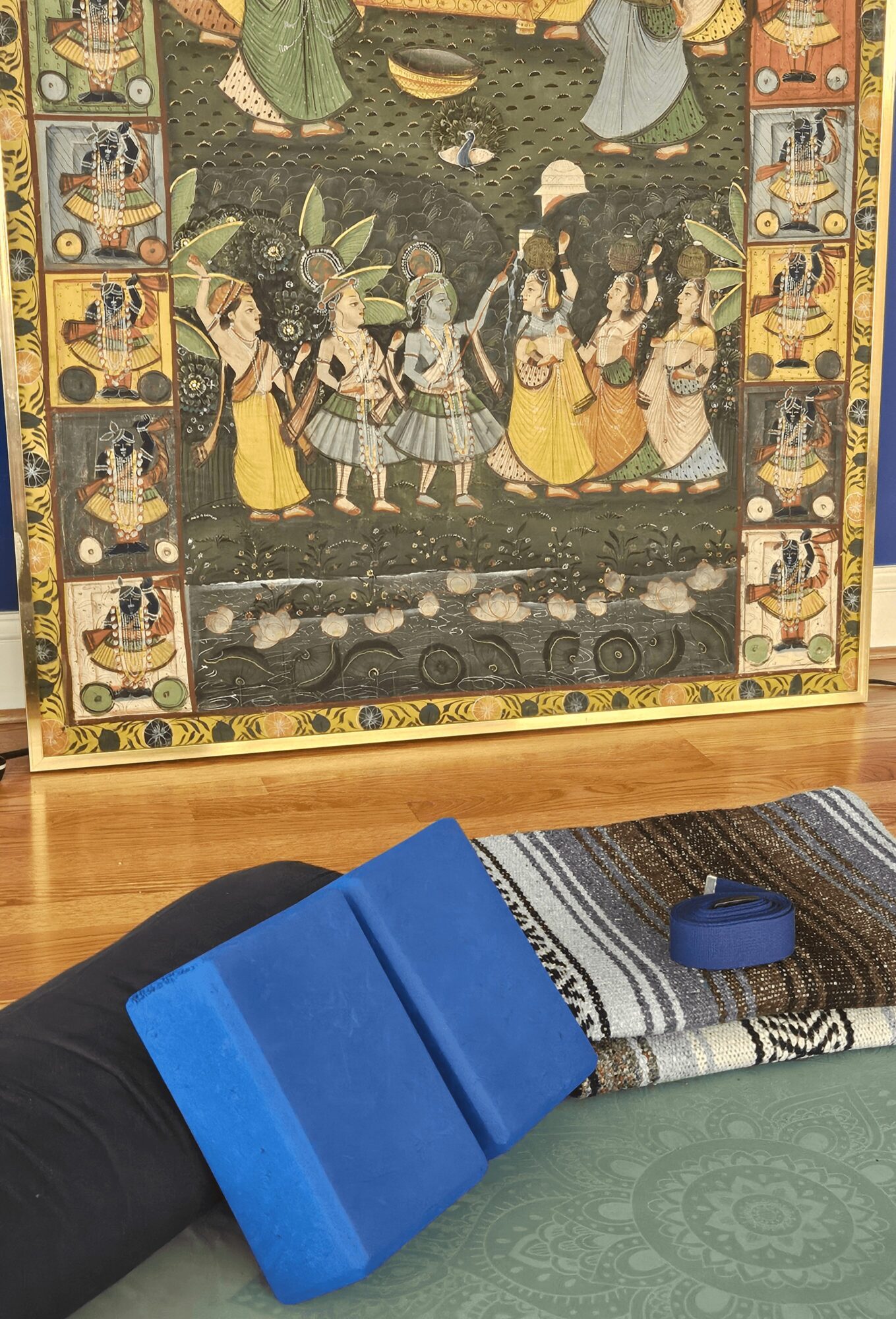Colorful painting depicting a group of people, framed and displayed on a wall, with a blue object and fabric in the foreground.
