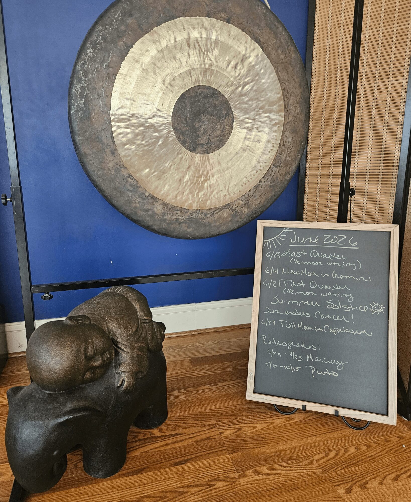 Large circular artwork on wall, sculpture of a seated person on wooden floor, chalkboard with handwritten notes nearby.