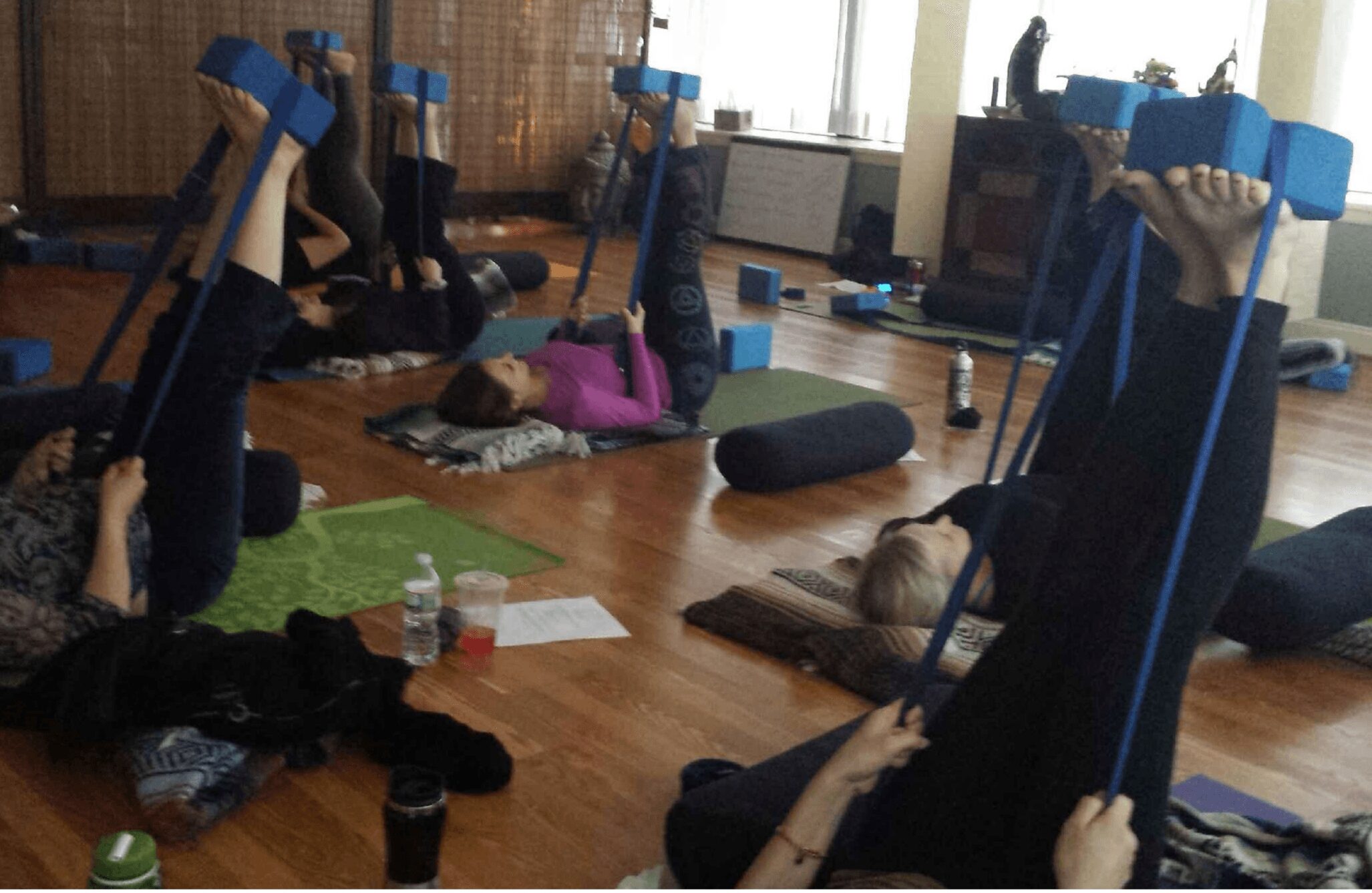 People lying on mats and holding resistance bands during a fitness class in a room with wooden floors and windows.