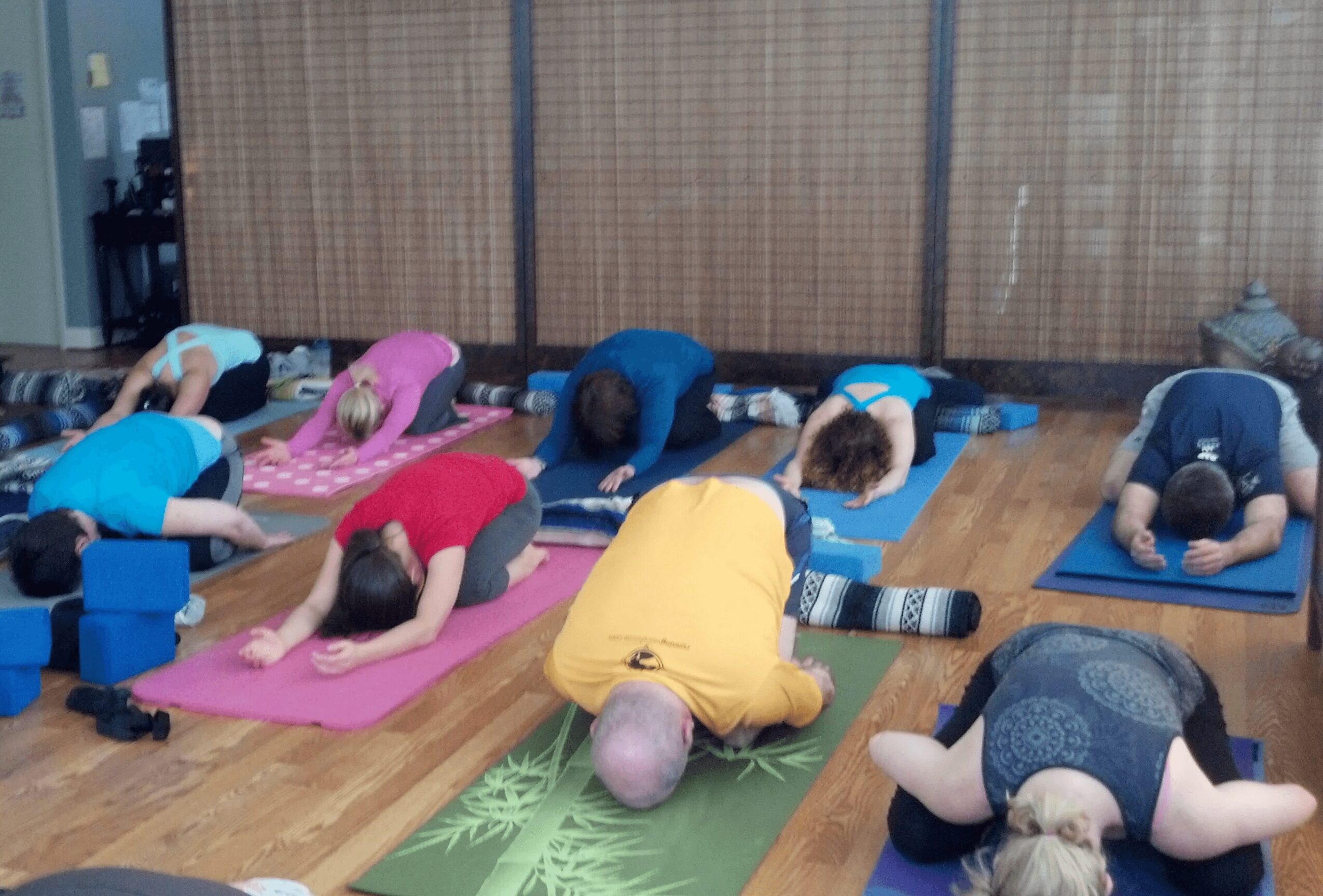 Group of people practicing yoga or meditation on mats in a room with wooden floor and bamboo wall, some with heads bowed.