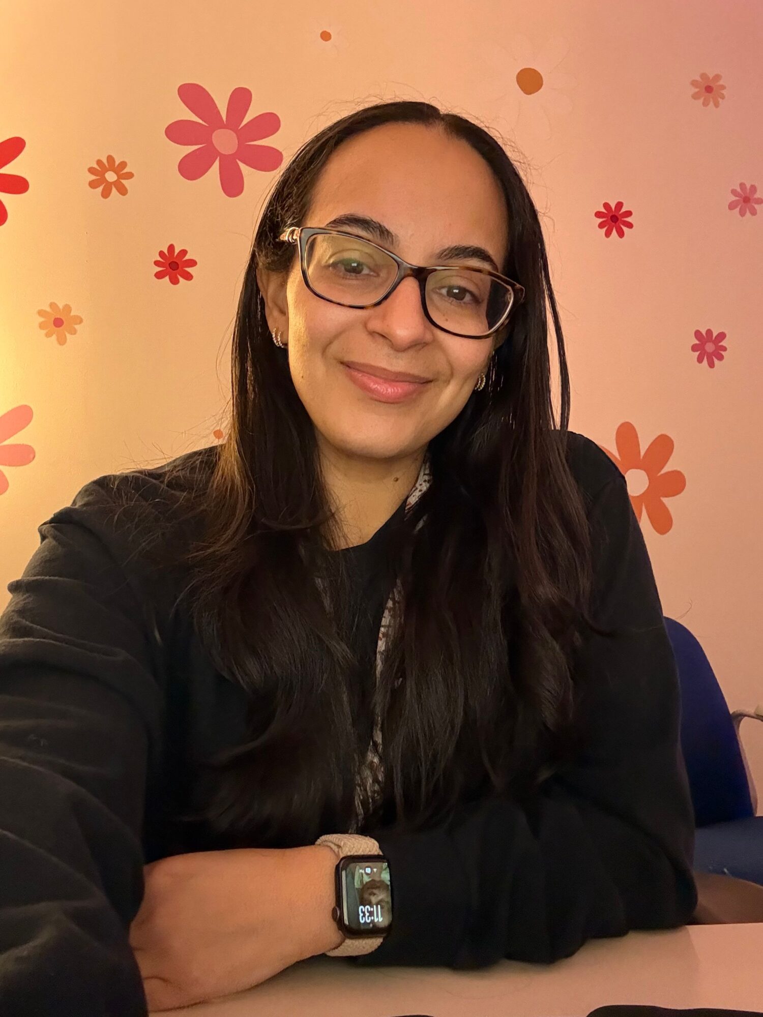 Woman with glasses smiling, long dark hair, wearing a black top, sitting at a table with pink and red flower decorations in background.