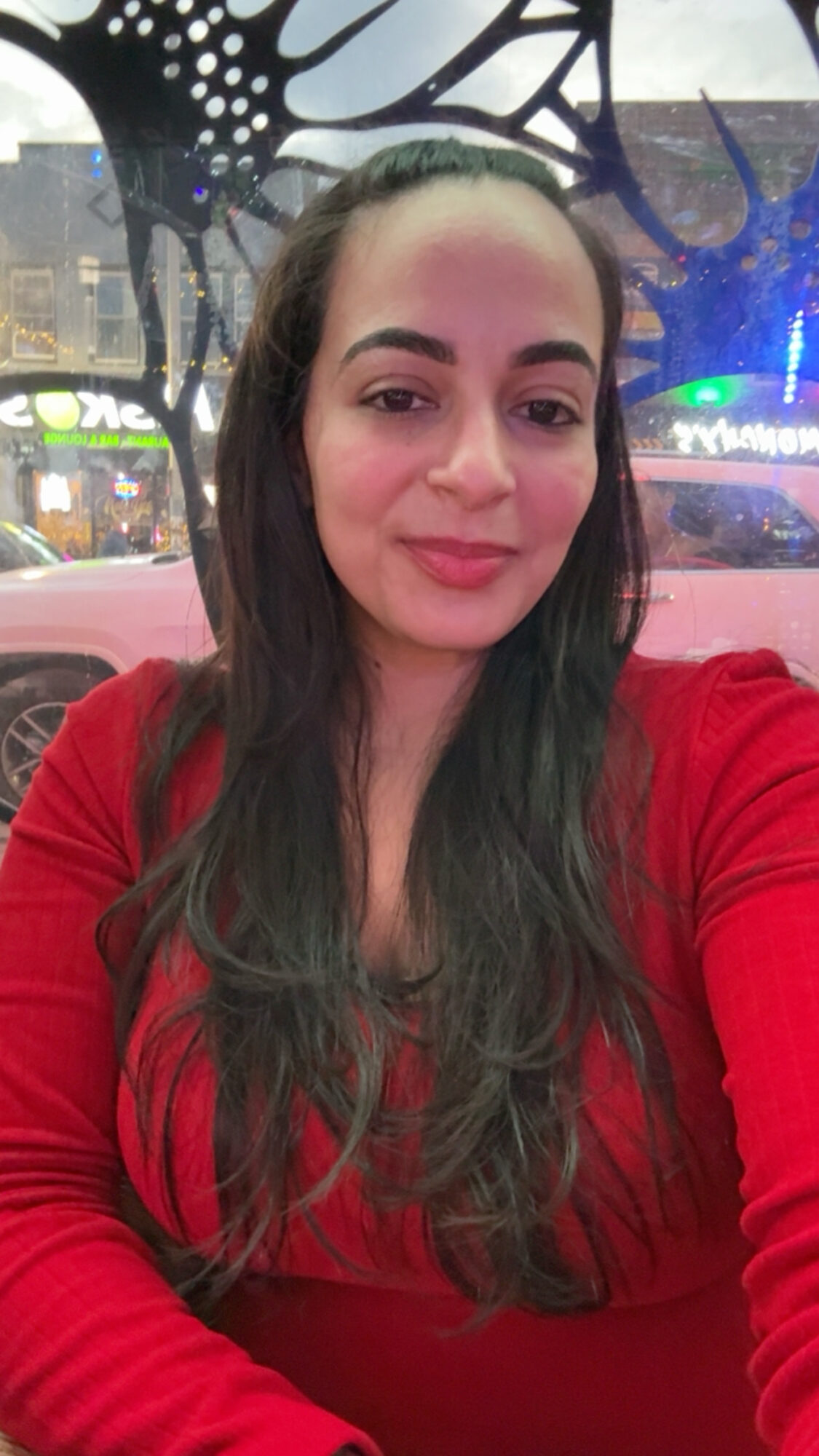 Woman with long dark hair wearing a red top, smiling, in front of a decorative background.