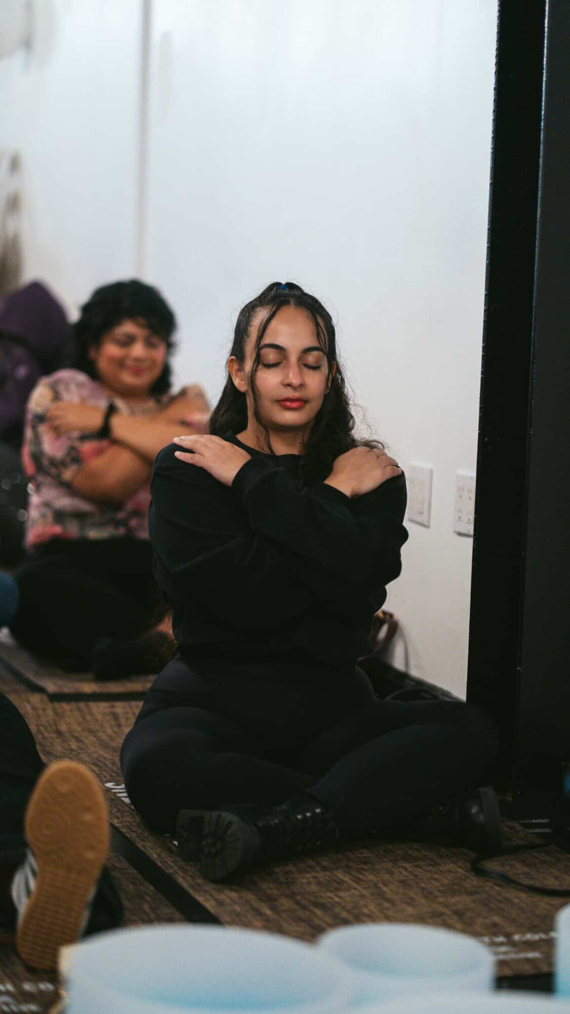Two women sitting on the floor, one in the foreground with crossed arms and the other in the background smiling.