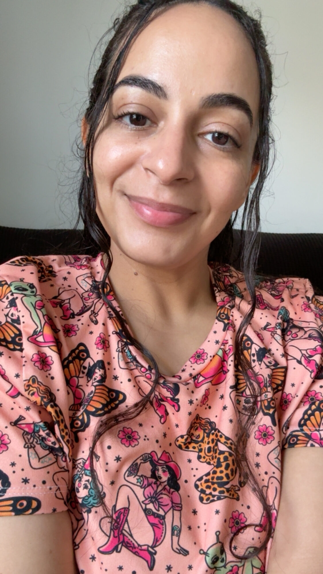 Young woman with dark hair, smiling, wearing a pink shirt with animal and floral patterns, sitting indoors.