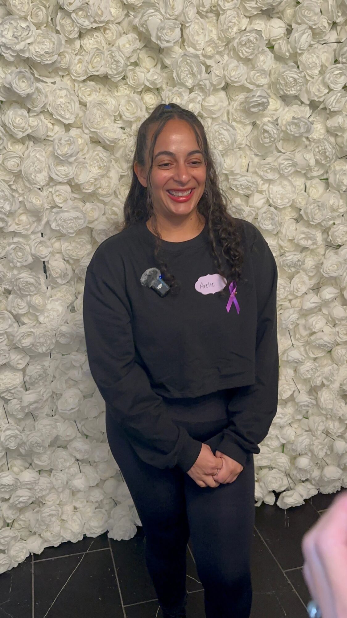 Woman with long curly hair smiling, wearing a black sweatshirt with cloud designs, standing in front of a white floral wall.