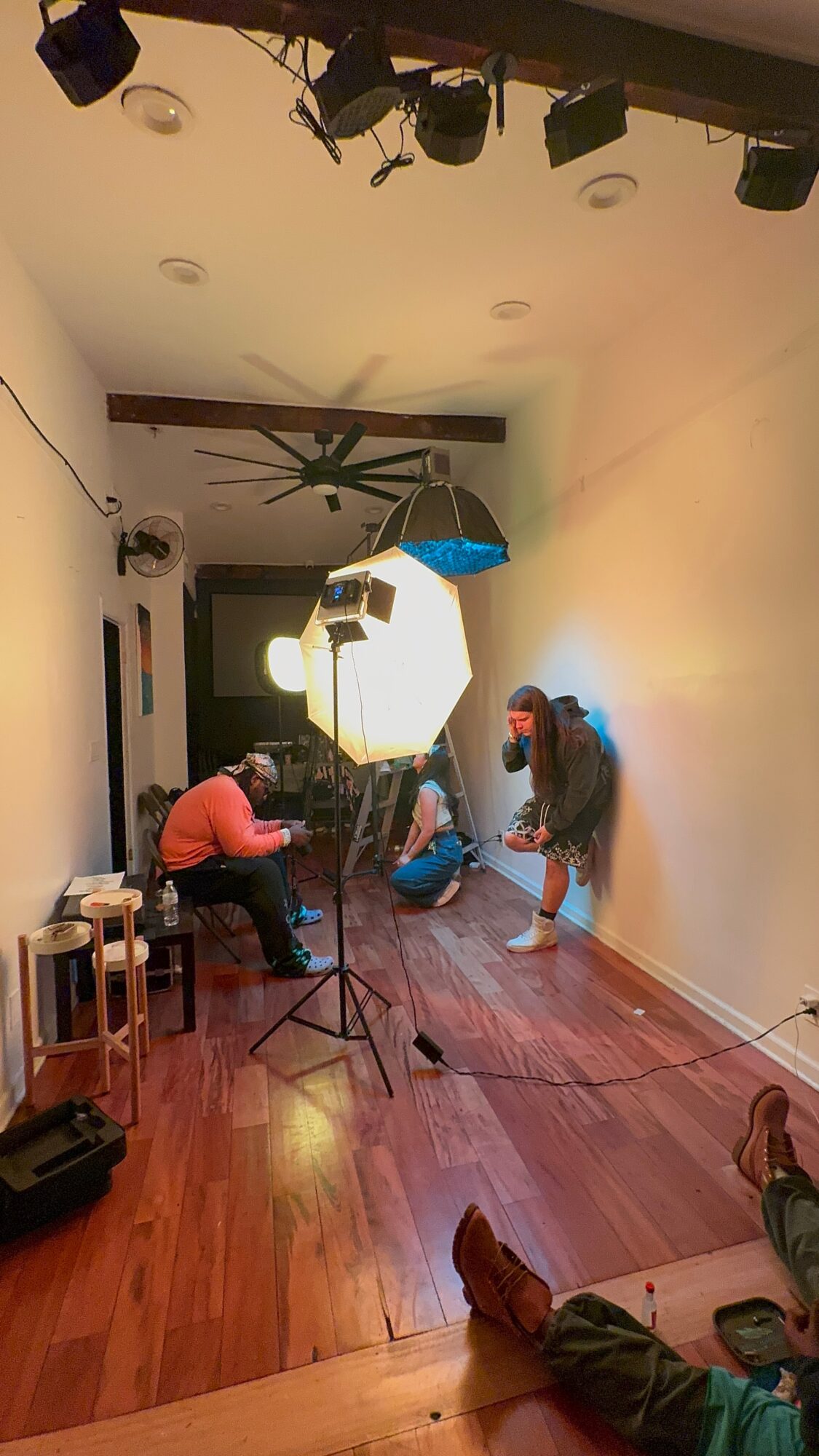 People in a room with wooden floor, some sitting and some standing, with lighting equipment and a large softbox, ceiling fan, and wall decor.