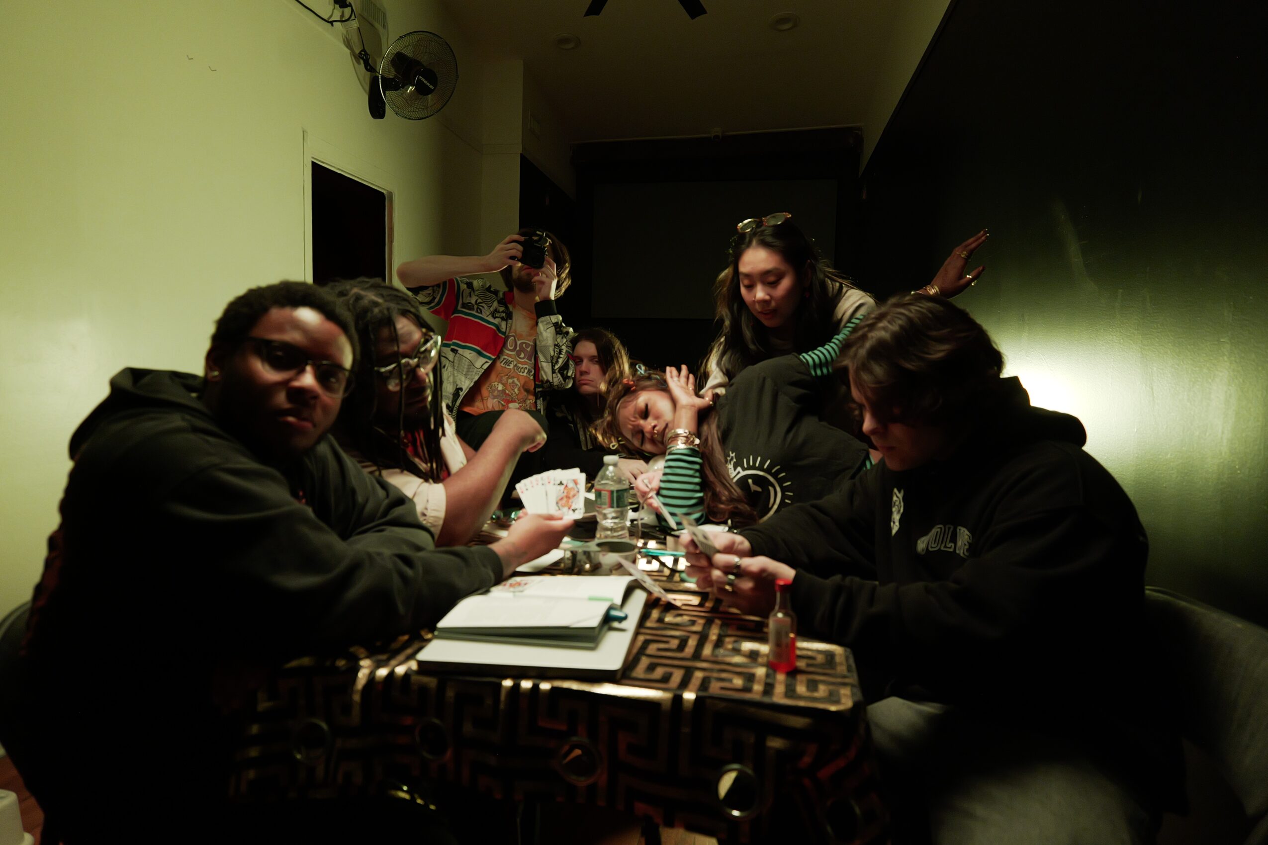 Group of people sitting around a table in a dimly lit room, some holding drinks and engaging with each other.
