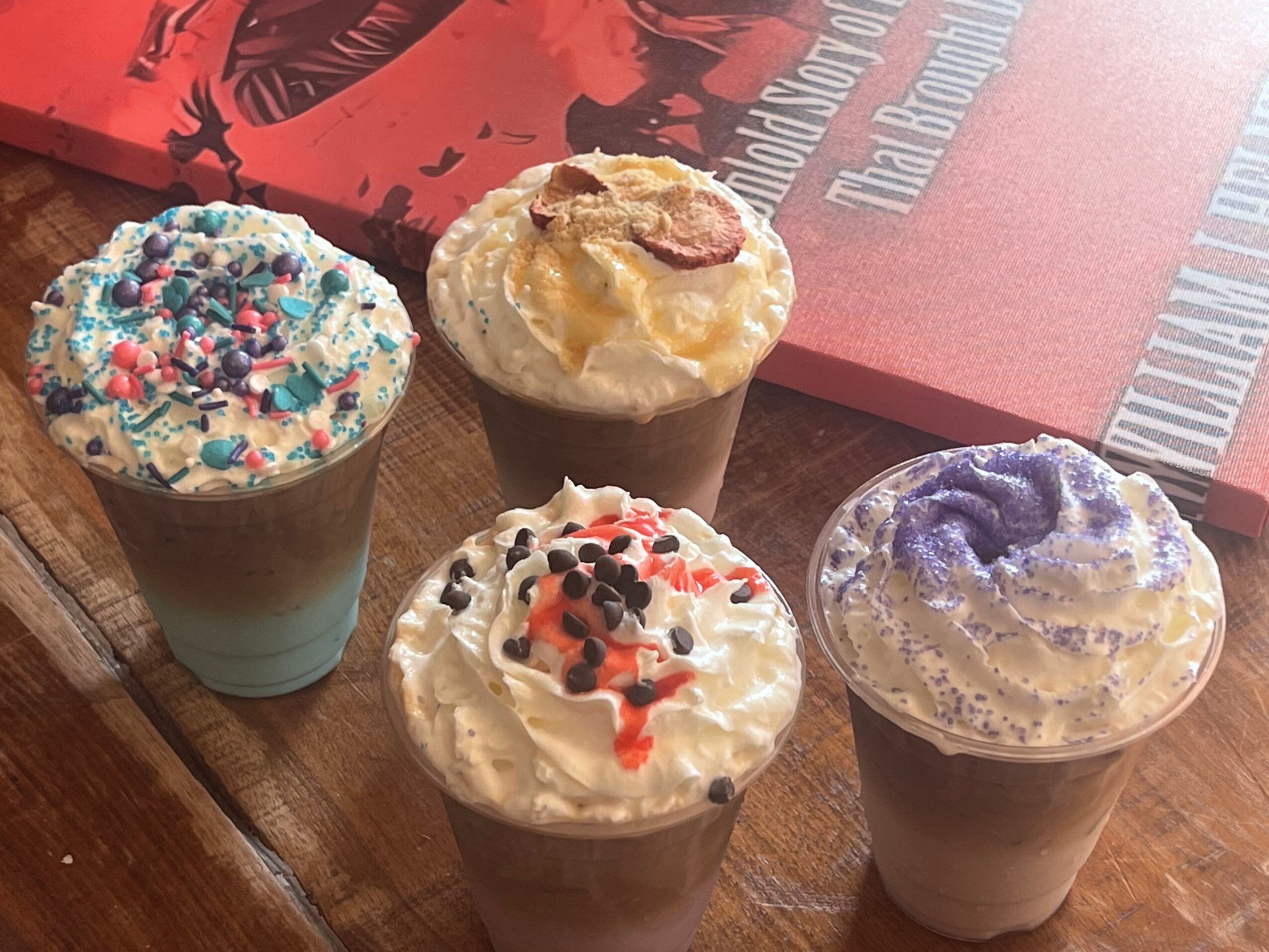 Four cupcakes with colorful toppings on a wooden surface, with a red book in the background.