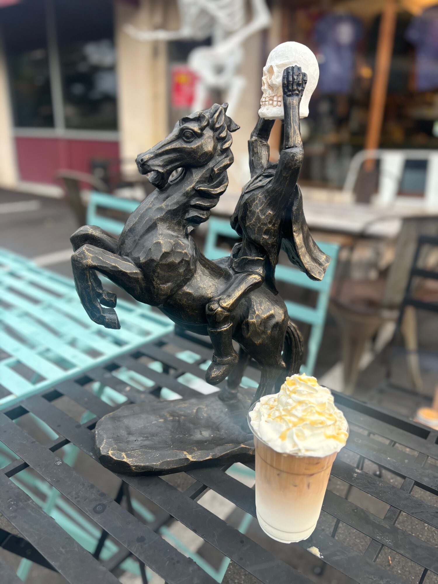 Statues of a horse and a person holding a skull, with a cup of ice cream on a table outdoors.