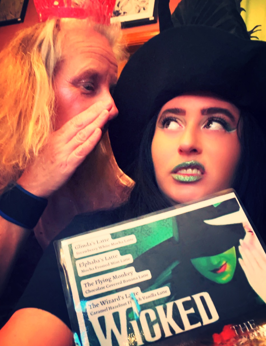 Two women talking, one with long blonde hair and the other with dark hair and a large black hat, holding a book titled 'WICKED'.