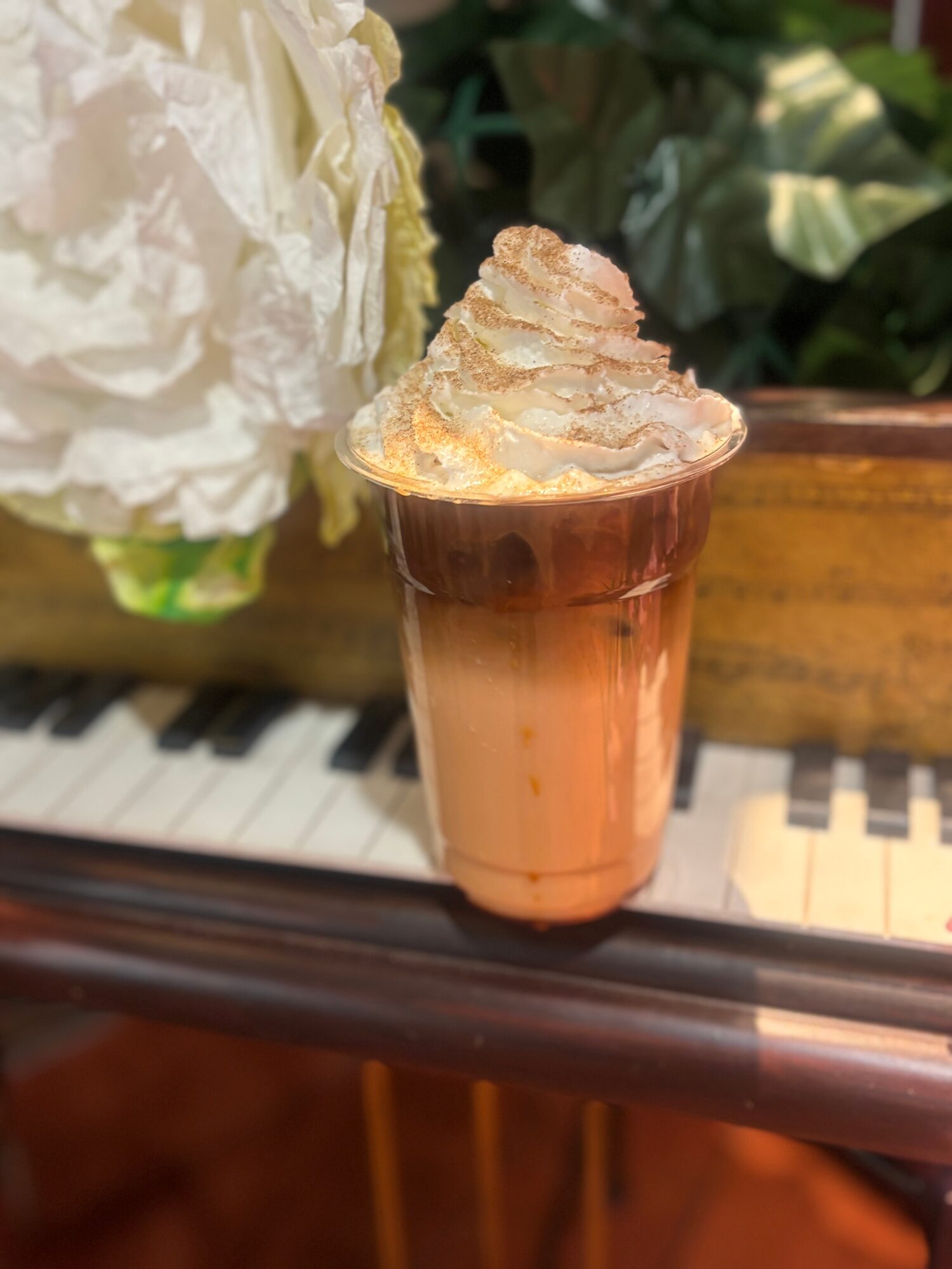 A tall glass of layered coffee drink topped with whipped cream, placed on a piano keyboard with plants in background.