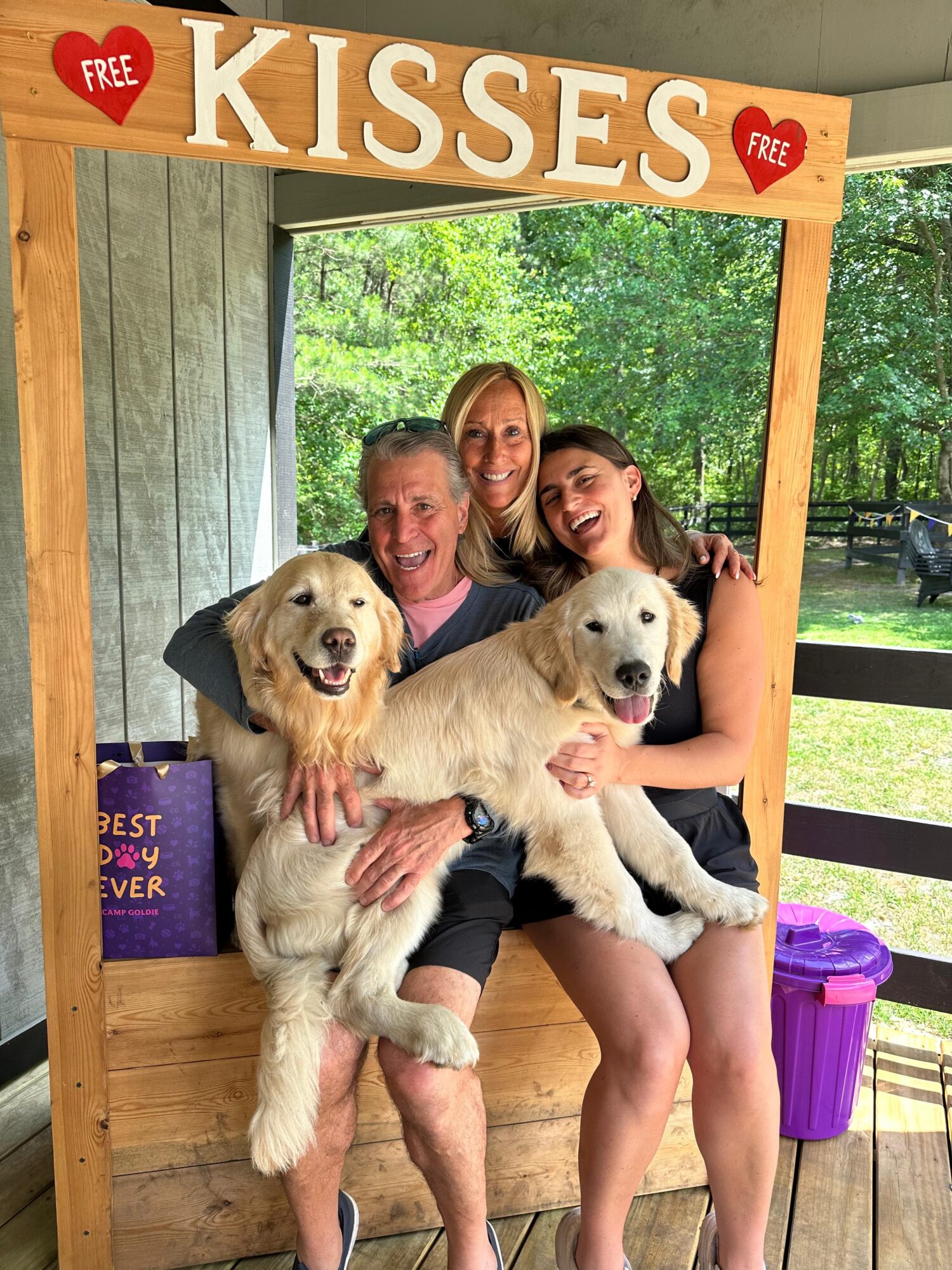 Three people smiling with two golden retriever puppies outdoors, under a wooden frame with a 'KISSES' sign, trees in background.