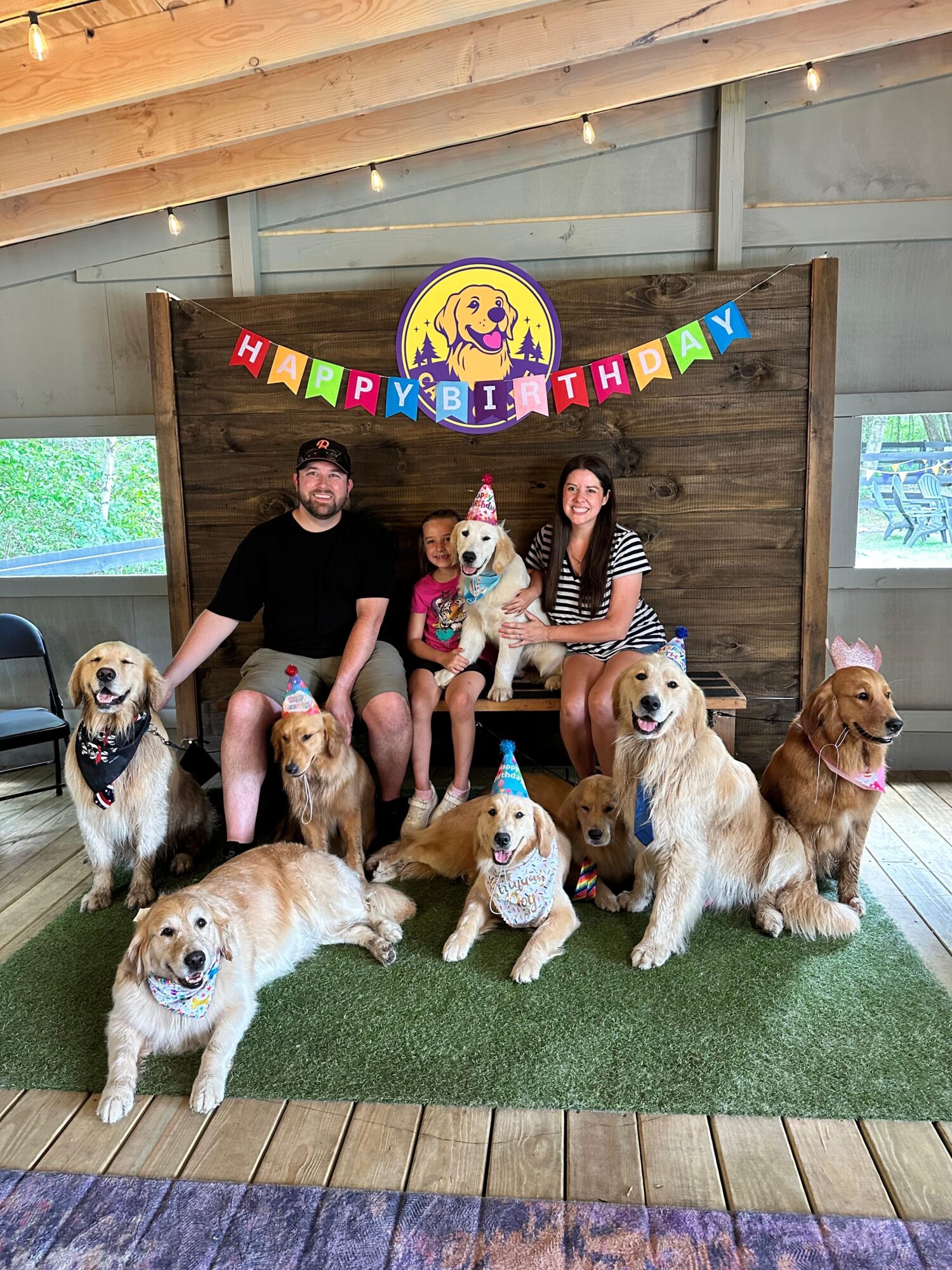 Two people and seven dogs sit on a bench with a birthday banner and a dog-themed sign behind them, inside a wooden enclosure.